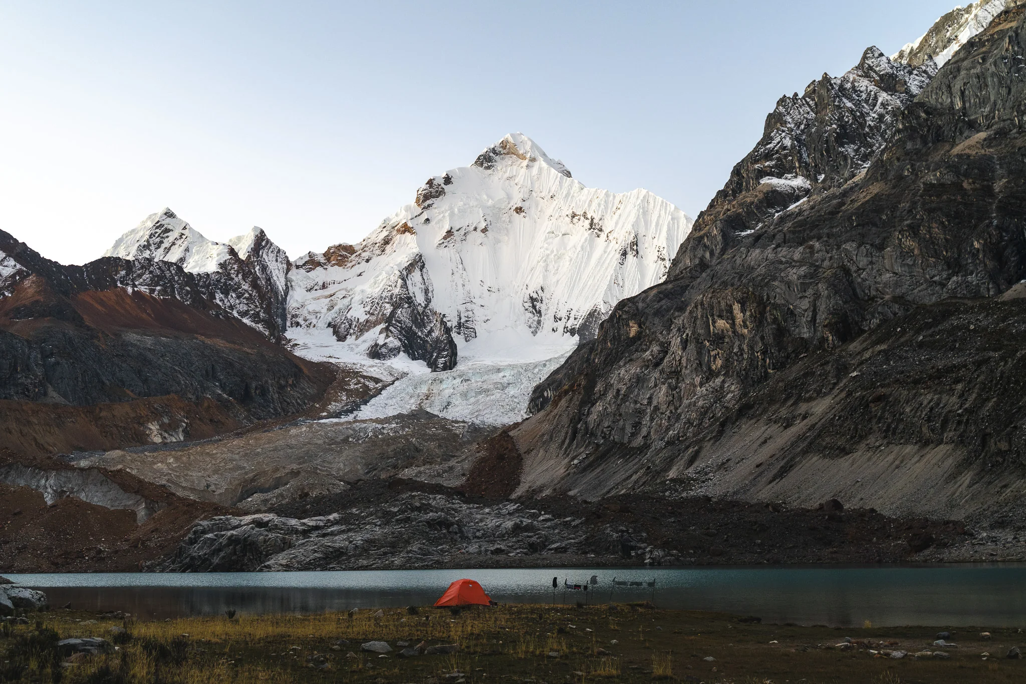 An orange tent on the shore of a still lake below a snow-covered peak and glacier, flanked by dark rock walls, Cordillera Huayhuash, Peru
