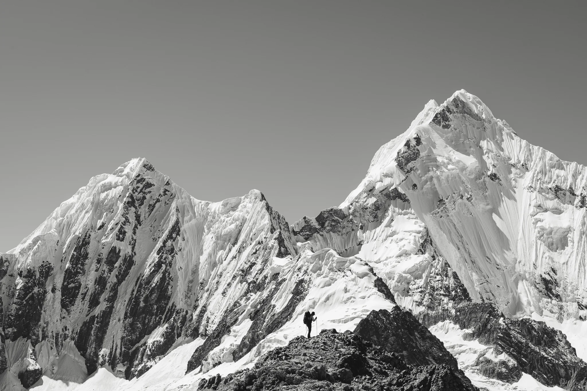 Black-and-white photo of a trekker standing on a dark ridge crest facing the snow-covered summits of Nevados Rasac and Yerupajá, Cordillera Huayhuash, Peru