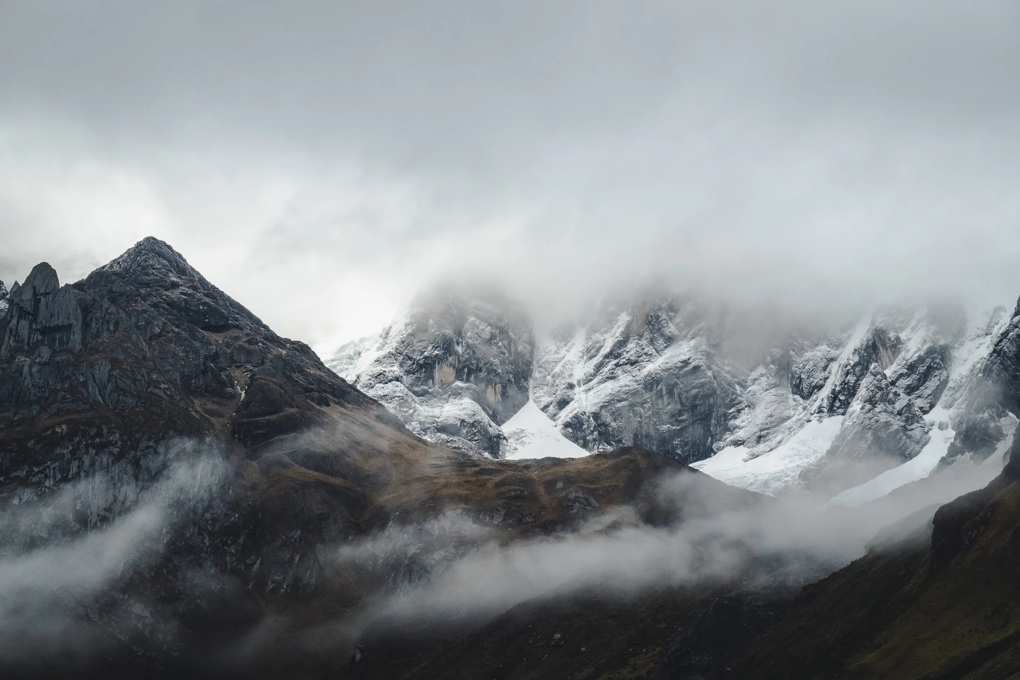 Cloud tearing across snow-covered peaks and dark rock ridges under a grey sky, east side of the Cordillera Huayhuash, Peru