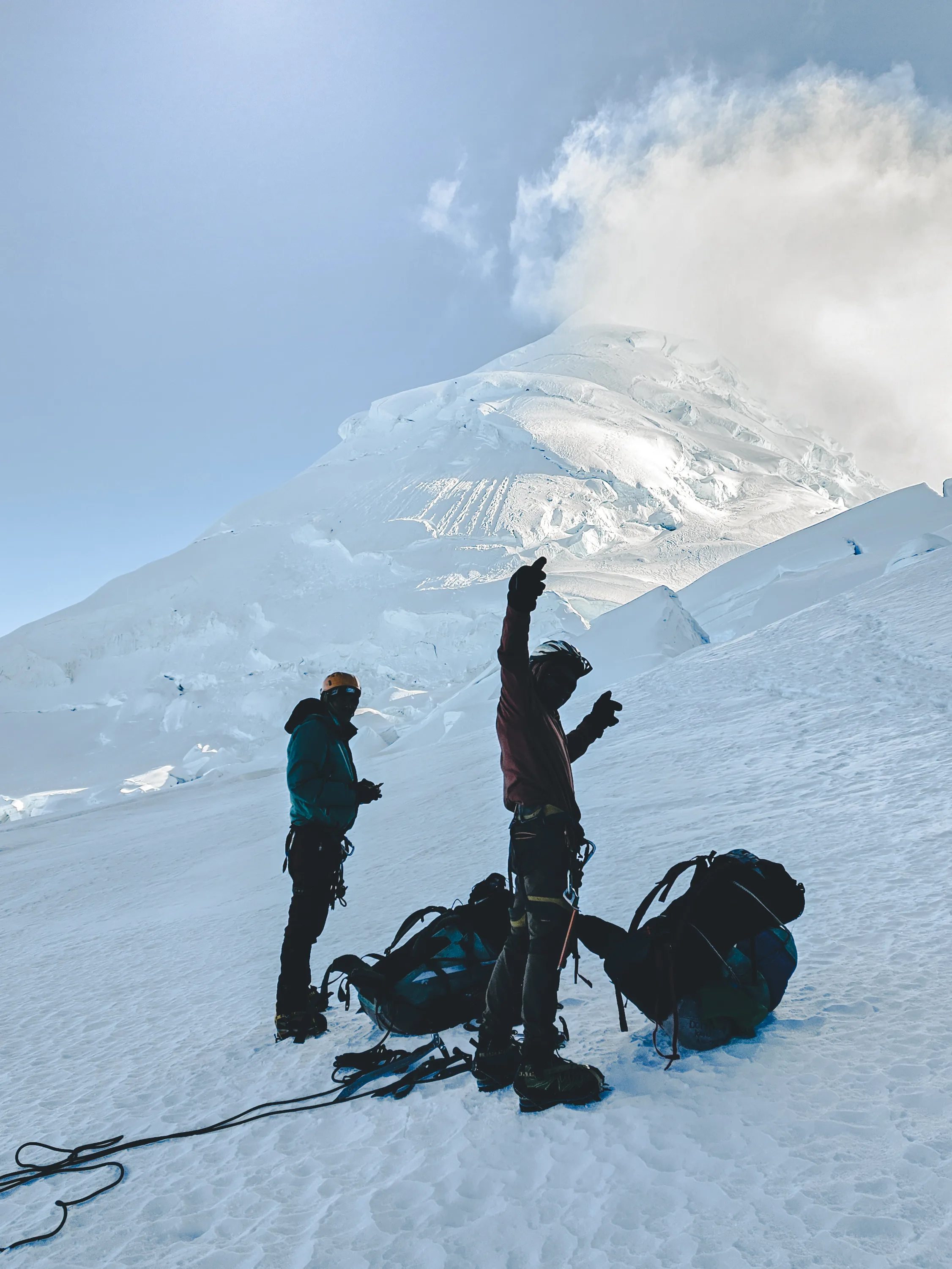 Vertical view of two climbers standing beside their packs on the glacier, one raising an arm, the snow dome of Nevado Huascarán and a cloud behind, Cordillera Blanca, Peru