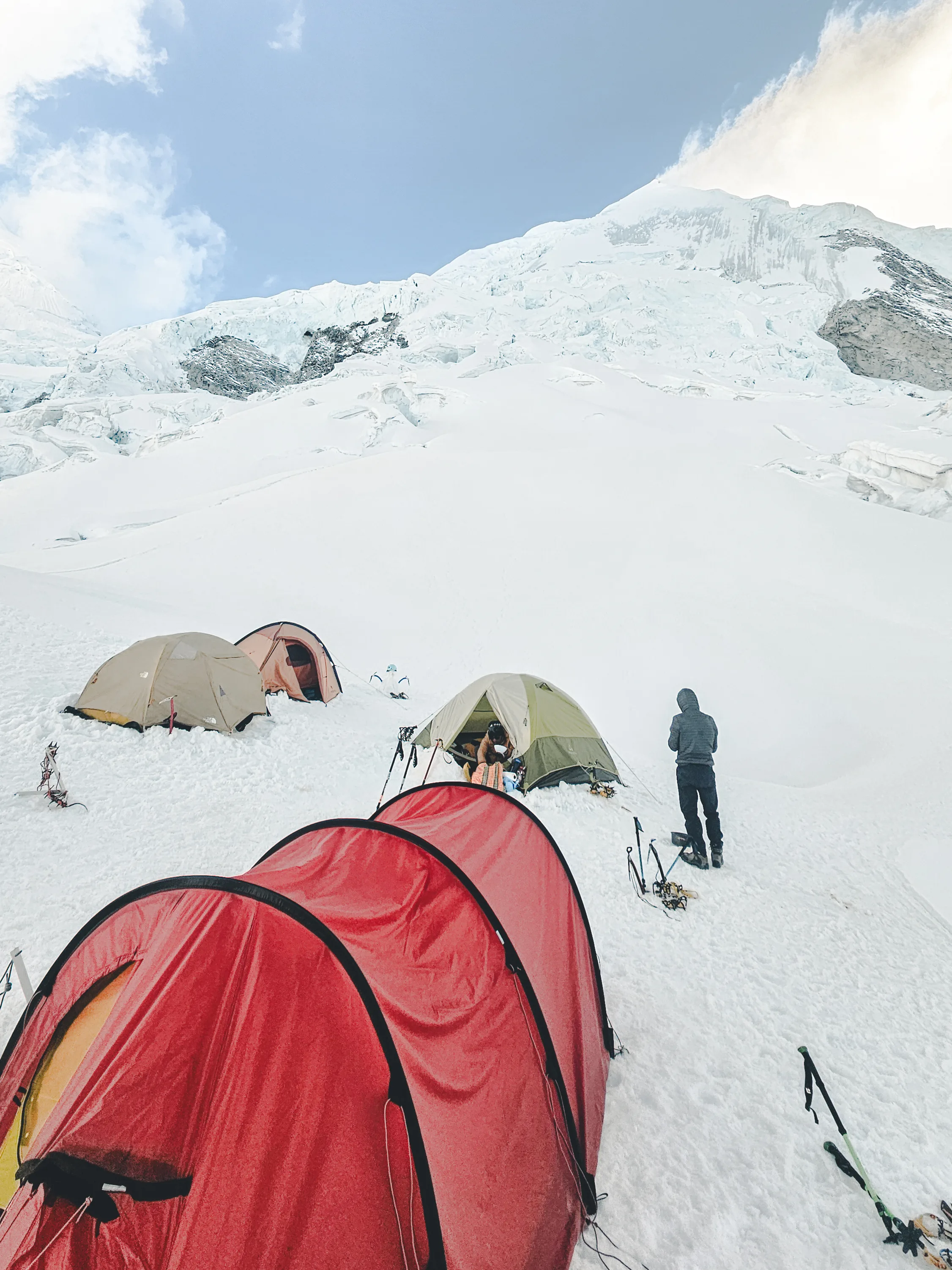 Vertical view of tents on the glacier at Camp 1 of Nevado Huascarán, a red tunnel tent in front and a climber standing by the tents behind, an icefall rising above, Cordillera Blanca, Peru