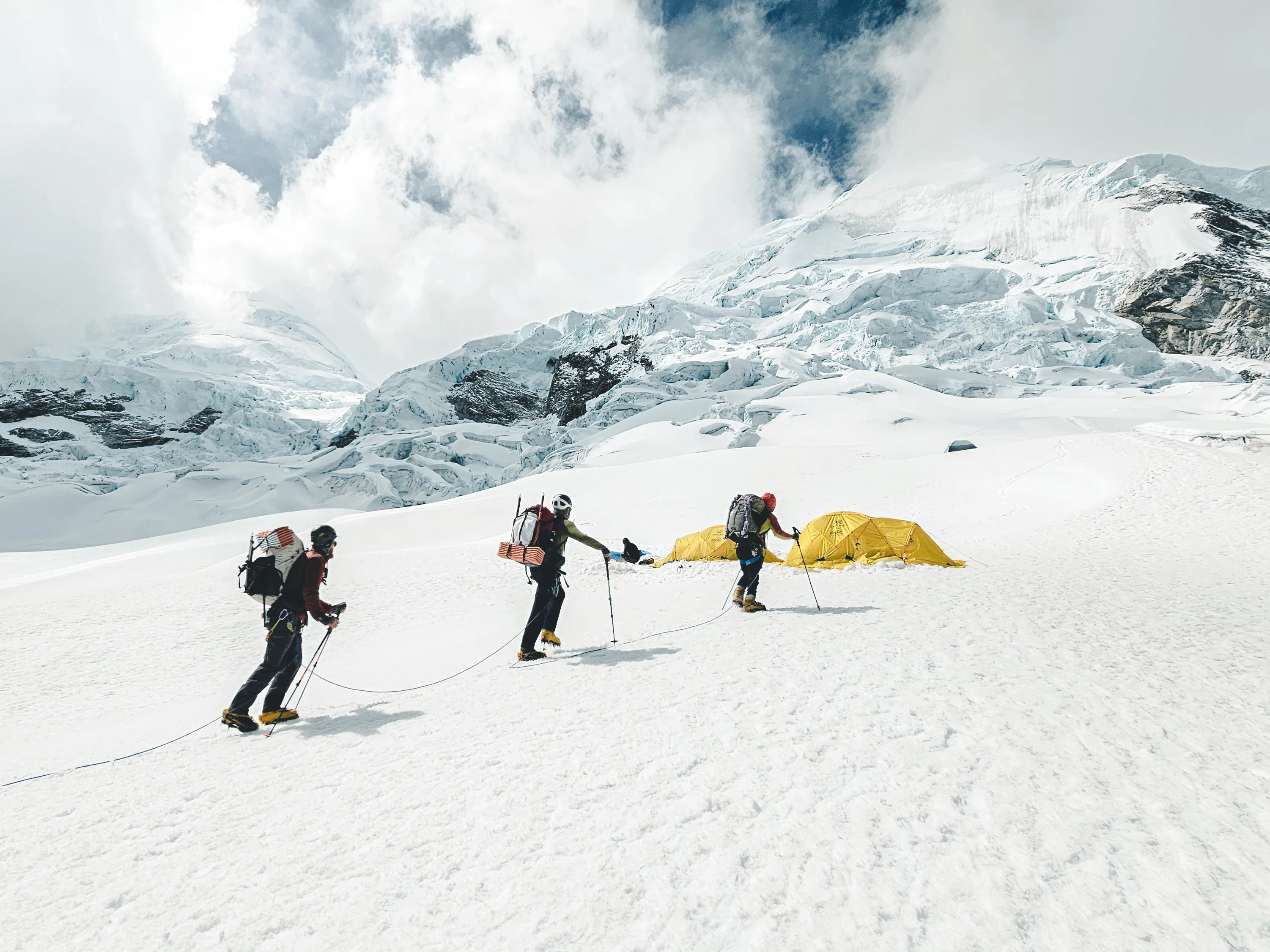 Three roped climbers with packs crossing the glacier toward yellow tents at Camp 1, the icefall of Nevado Huascarán and clouds behind, Cordillera Blanca, Peru