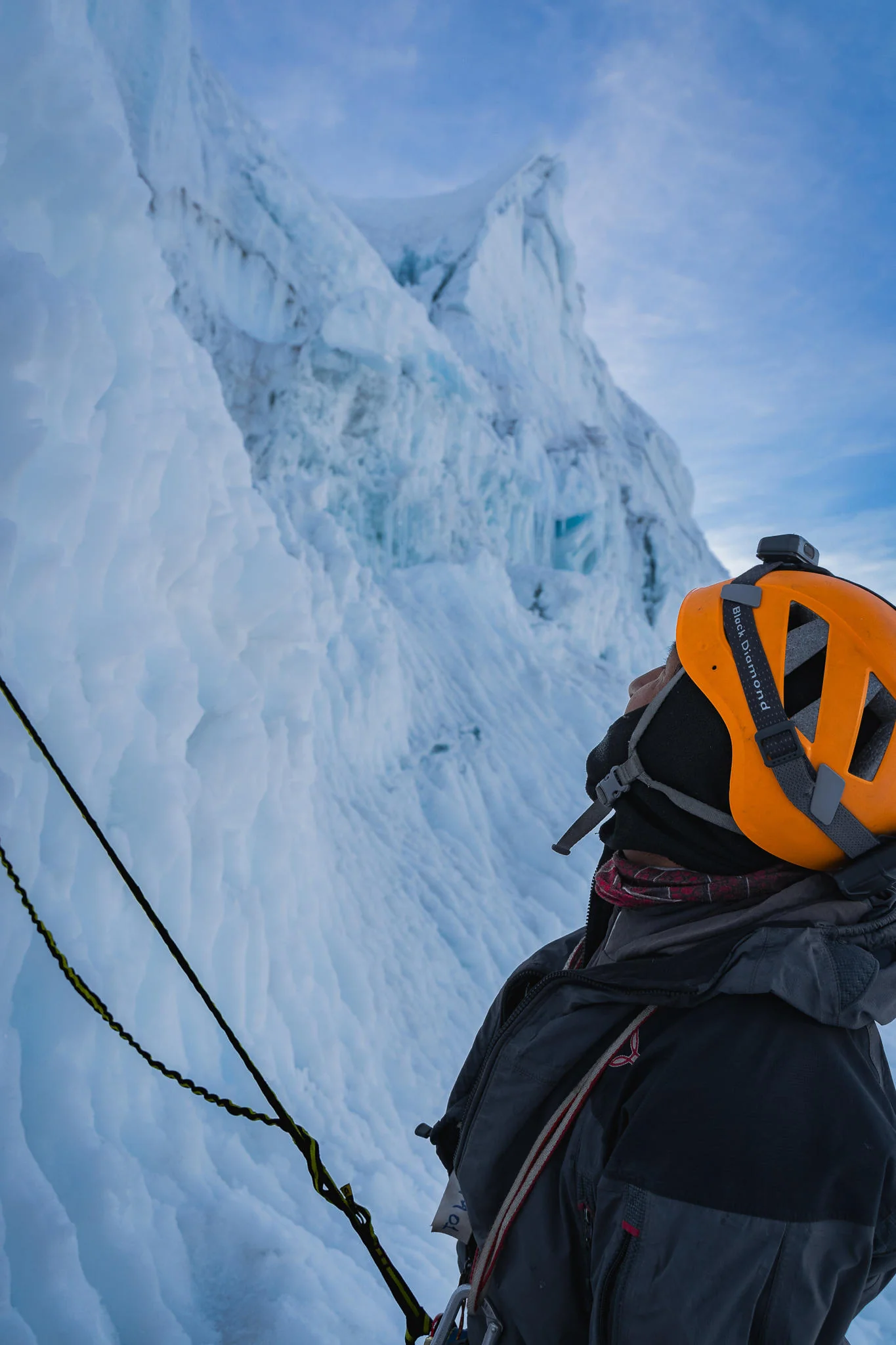 A climber in an orange helmet looking up at a wall of blue glacier ice, ropes running up the face, Nevado Yanapaccha, Huascarán National Park, Peru