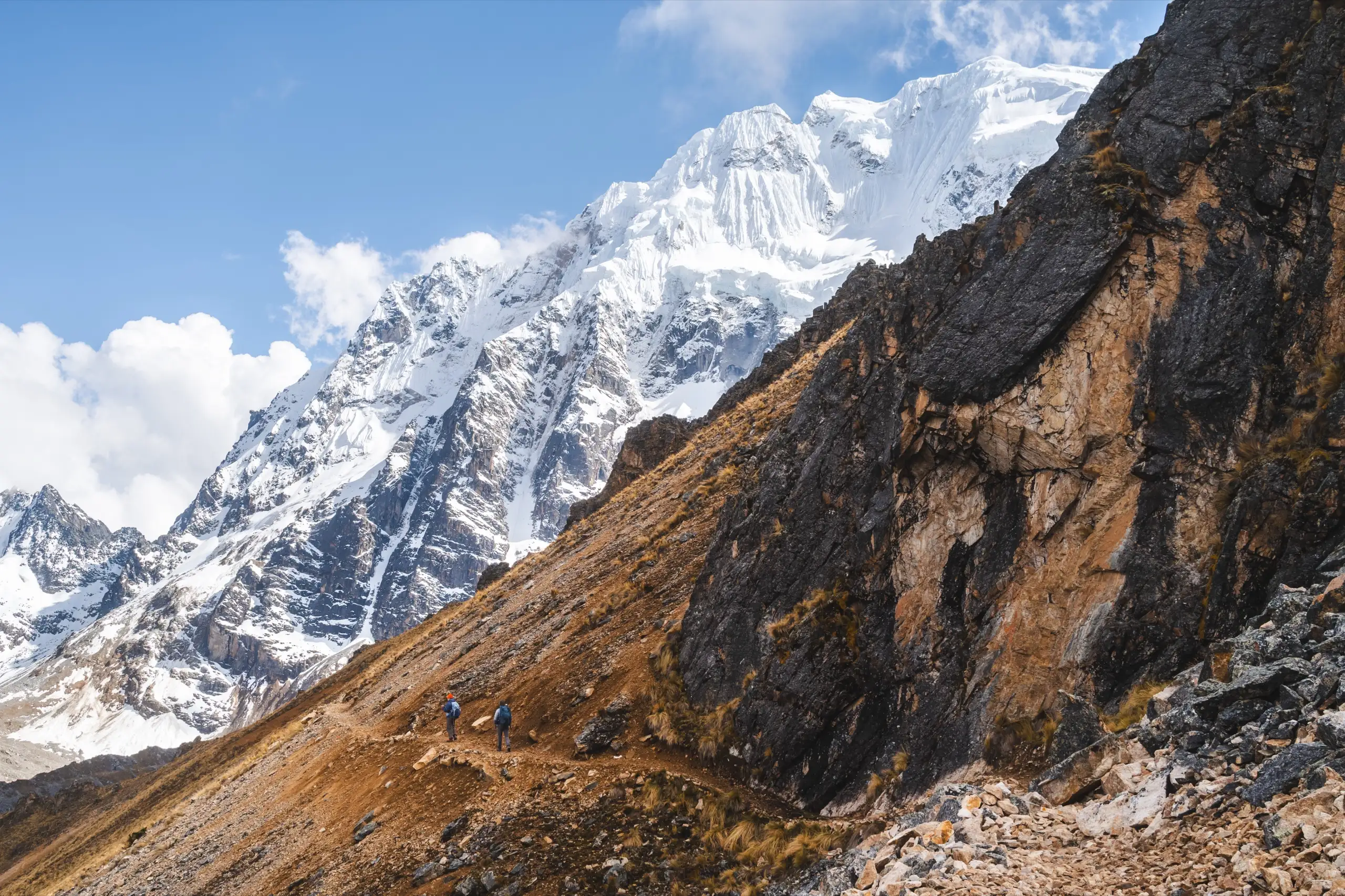 Small trekkers on a trail across a golden slope beneath the huge glaciated face of Nevado Salkantay and a dark rock cliff, Nevado Salkantay, Cordillera Vilcabamba, Peru