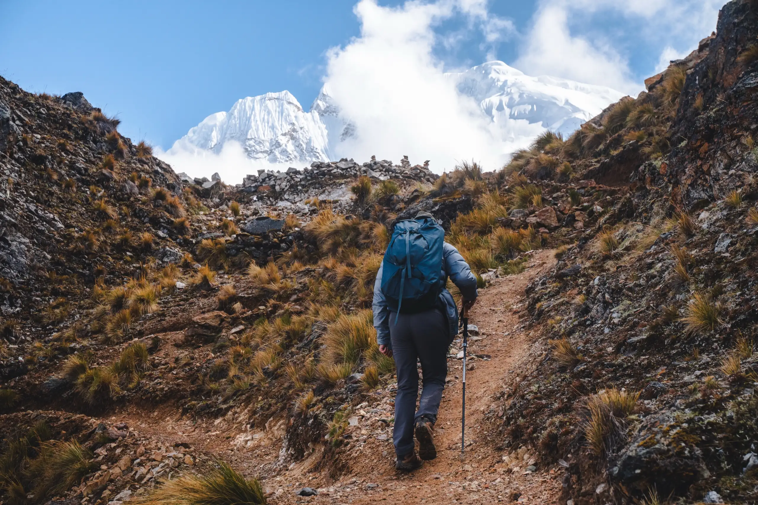 A trekker with a blue backpack and trekking pole climbing a rocky trail between brown slopes toward a snowy peak half hidden in cloud, Nevado Salkantay, Cordillera Vilcabamba, Peru
