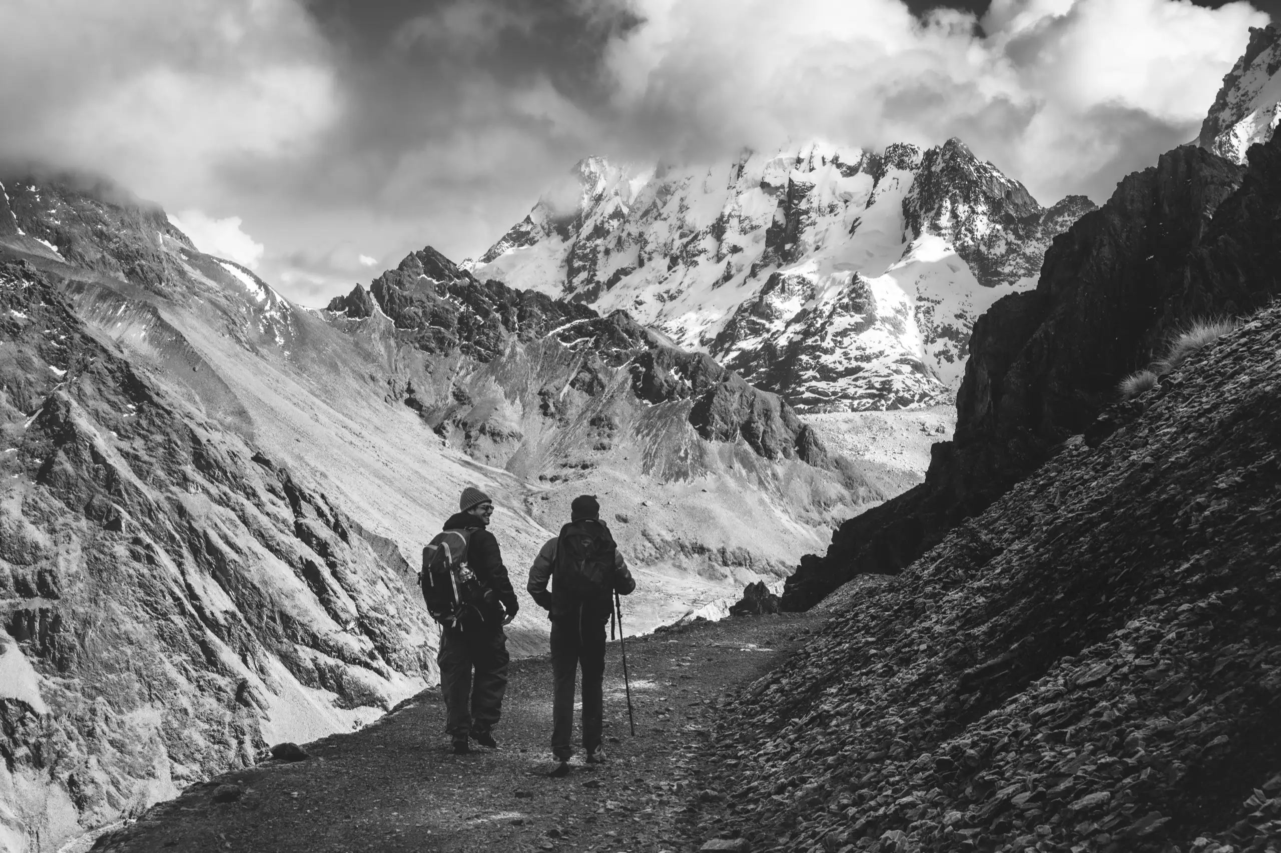Black-and-white photo of two trekkers walking along a trail cut into a steep slope, jagged snow-covered peaks and cloud ahead, Ahobamba Trek, Cusco region, Peru