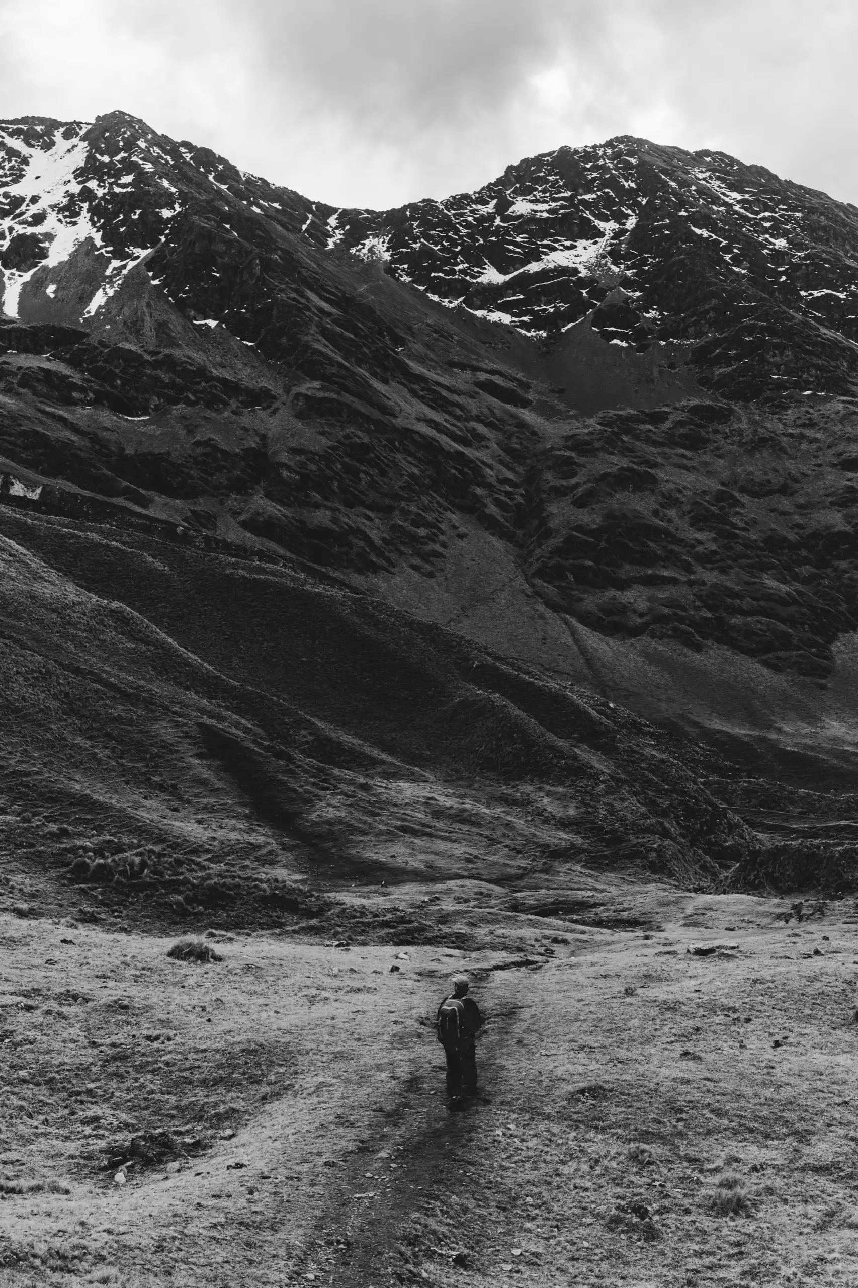 Vertical black-and-white photo of a lone trekker on a dirt trail in a wide valley, snow-streaked peaks and heavy cloud above, Ahobamba Trek, Cusco region, Peru