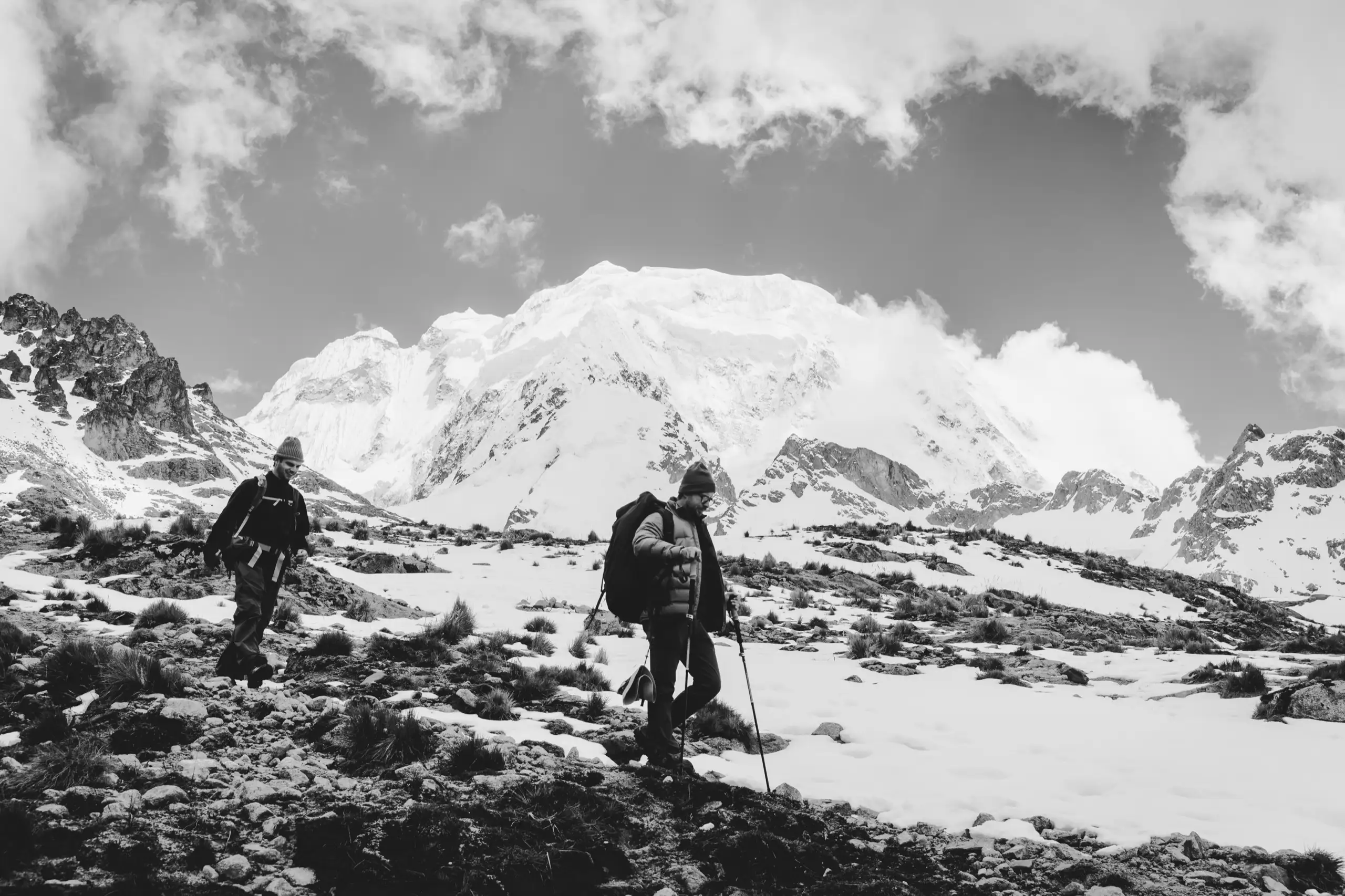 Black-and-white photo of two trekkers with poles crossing snowy, rocky ground, the huge snow-covered mass of Nevado Salkantay and cloud behind them, Nevado Salkantay, Cordillera Vilcabamba, Peru
