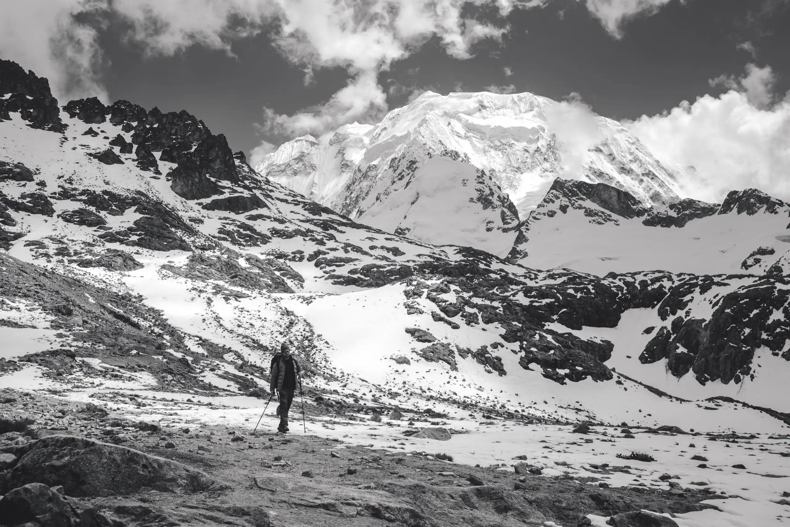Black-and-white photo of a lone trekker on a snowy, rocky slope beneath the broad snow-covered east face of Nevado Salkantay, Nevado Salkantay, Cordillera Vilcabamba, Peru