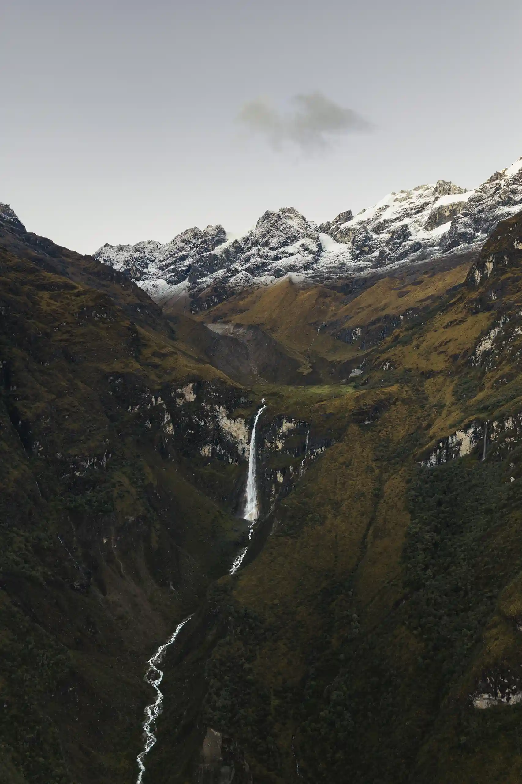 A waterfall streaming down a steep green valley wall beneath a line of snow-covered peaks, its river winding along the valley floor below, Ahobamba Valley, Peru