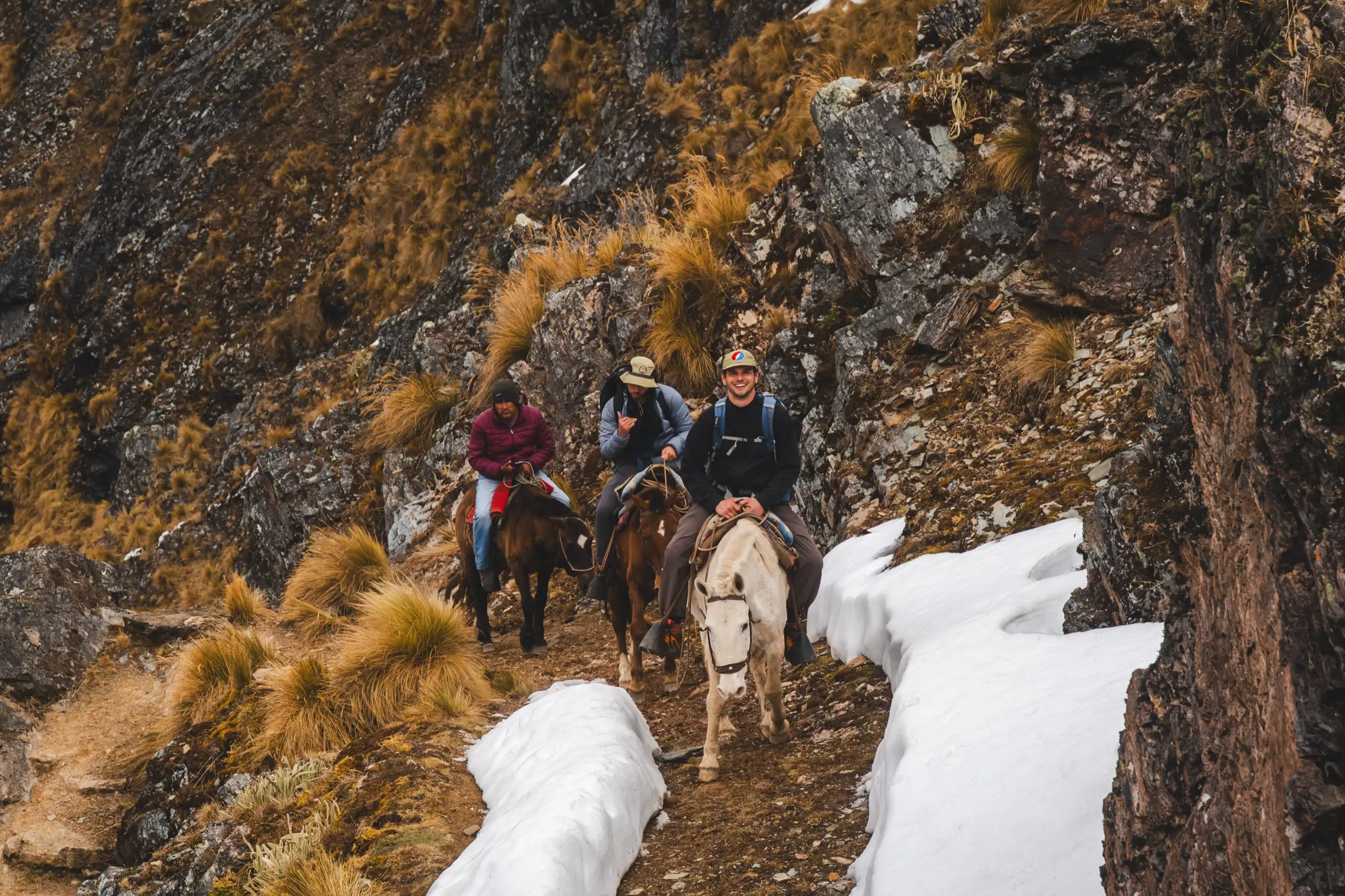 Three smiling riders on horses on a rocky trail between banks of snow, golden grass and a steep rock wall around them, Ahobamba Trek, Cusco region, Peru