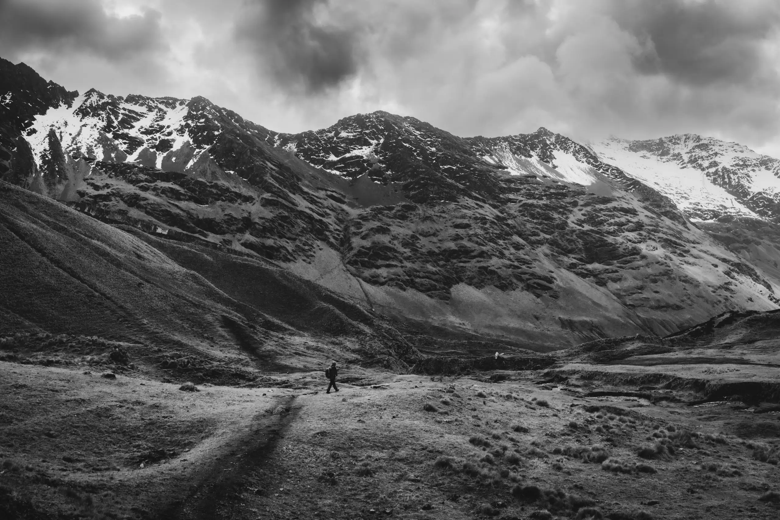 Black-and-white photo of a lone trekker on a trail across a wide high valley, snow-dusted mountains and heavy cloud above, Ahobamba Trek, Cusco region, Peru