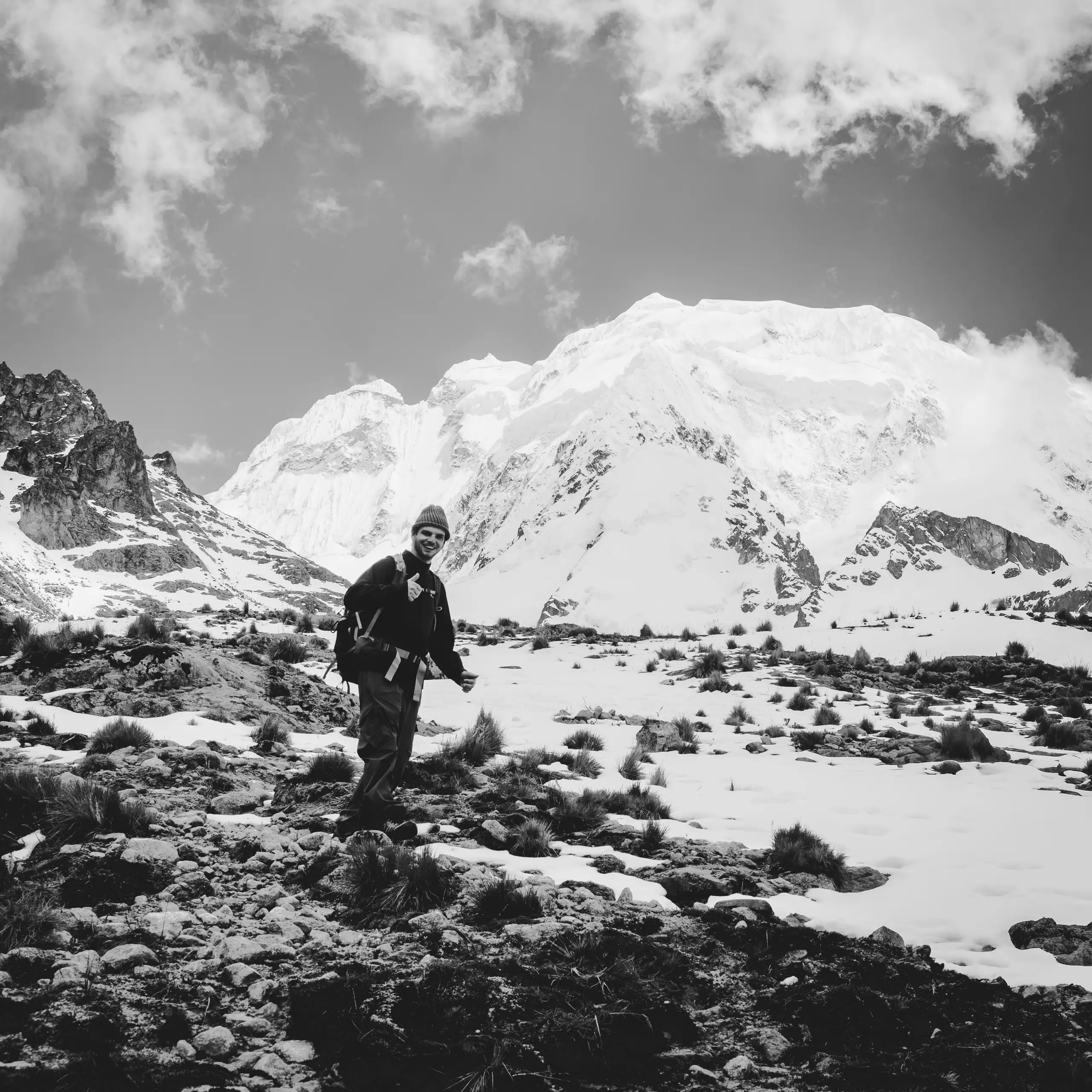 Black-and-white photo of a trekker standing among snowy rocks, the snow-covered bulk of Nevado Salkantay rising behind under broken cloud, Nevado Salkantay, Cordillera Vilcabamba, Peru