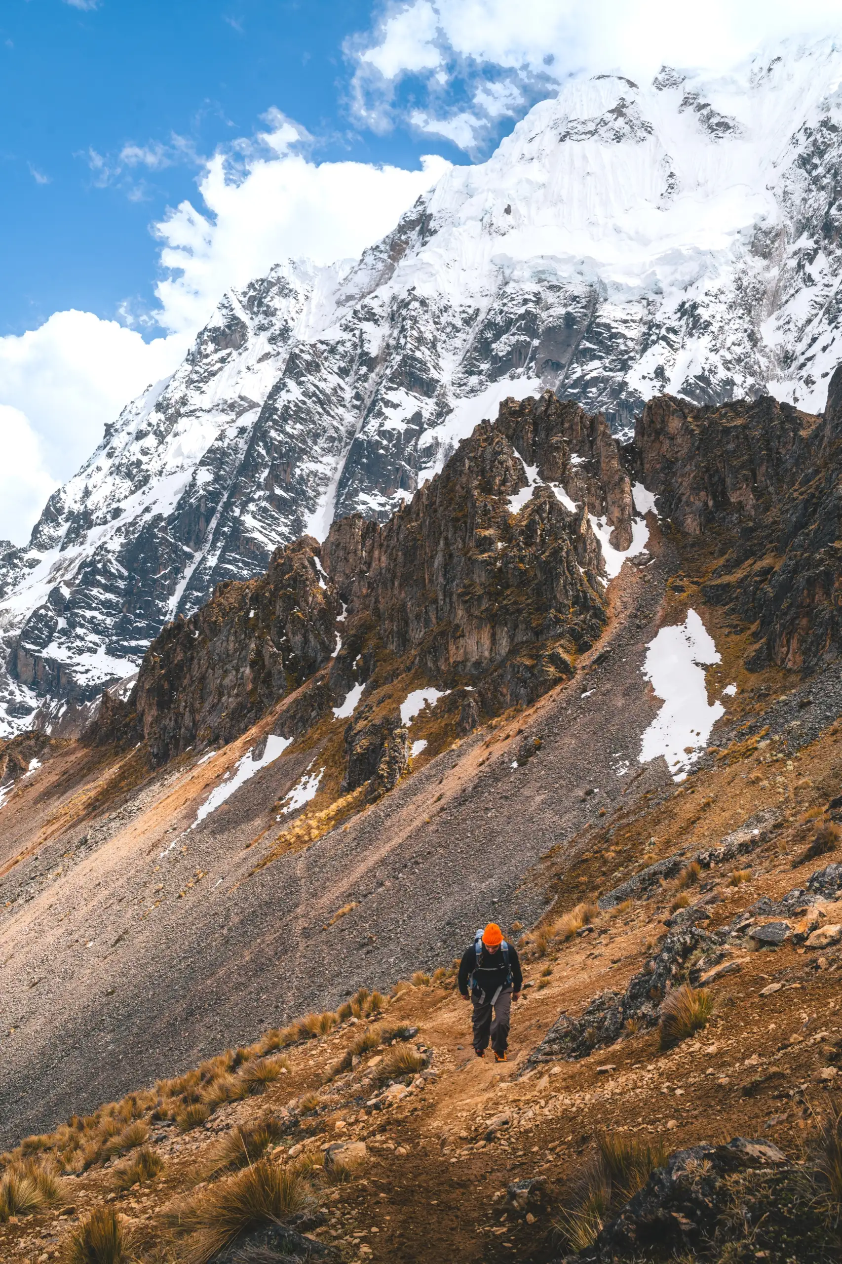 Vertical view of a trekker in an orange hat climbing a brown grassy slope below a jagged snow-covered peak and blue sky, Nevado Salkantay, Cordillera Vilcabamba, Peru