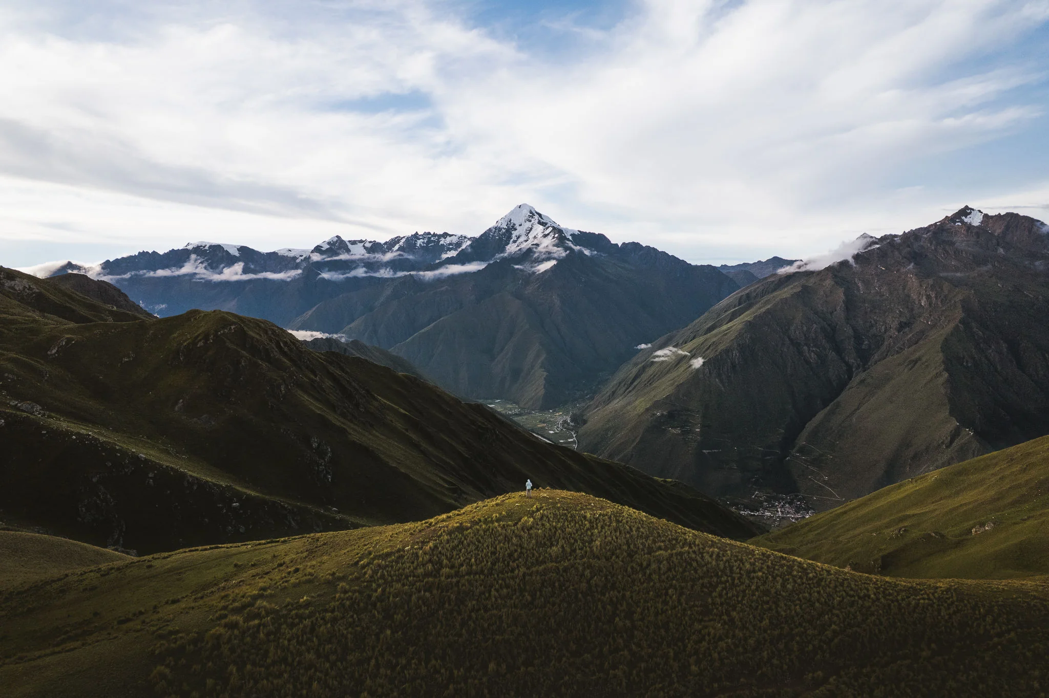 A lone hiker on a broad grassy ridge above a deep valley, a snow-capped peak rising beyond under a bright sky, Ancascocha, Sacred Valley, Peru