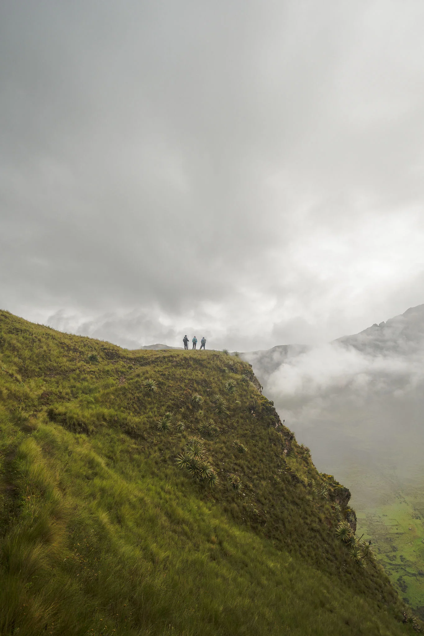 Three trekkers standing on the edge of a grassy ridge under heavy cloud, mist rising from the valley below, above Ollantaytambo, Sacred Valley, Peru