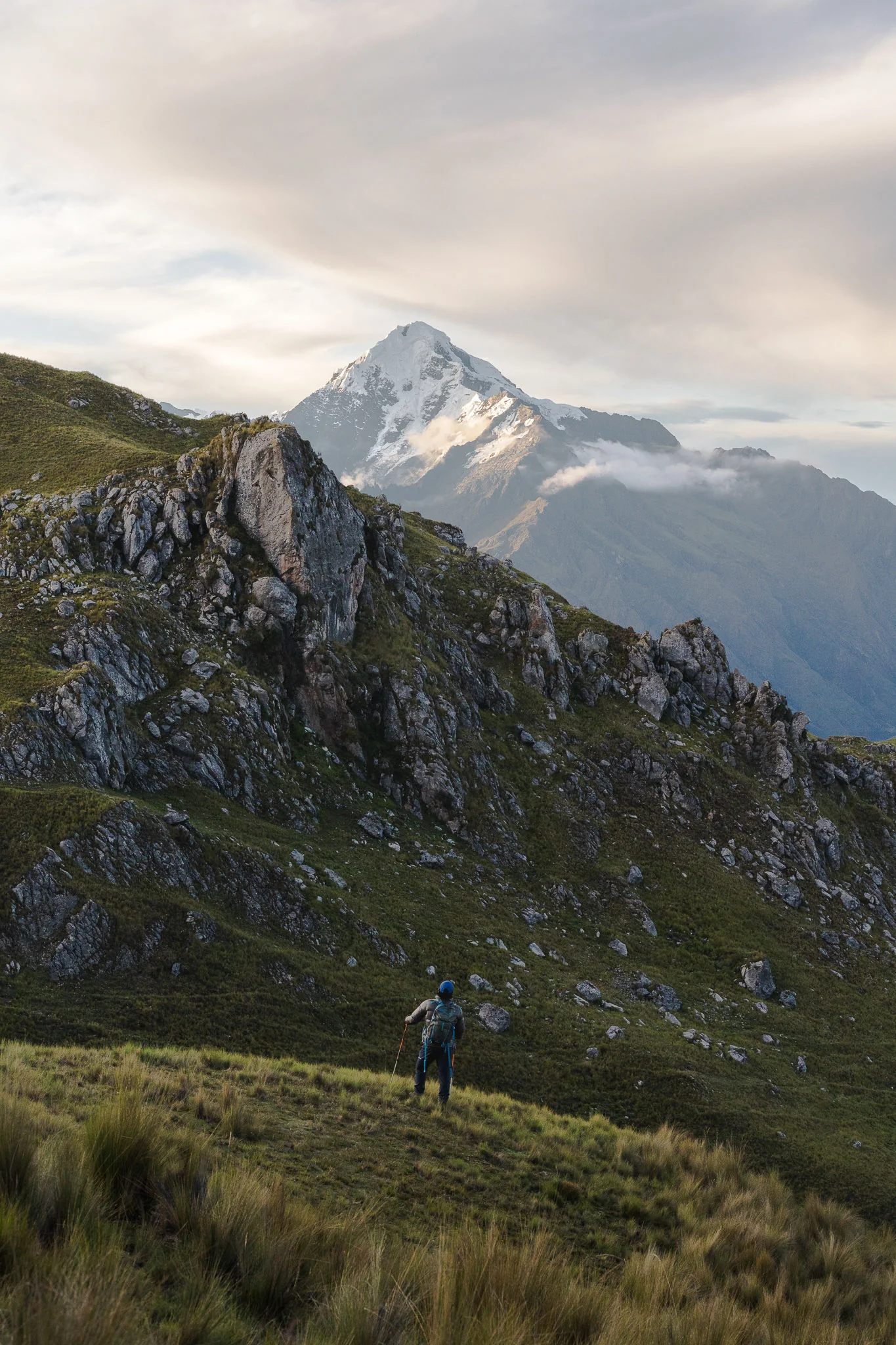 A trekker walking up a grassy slope past rocky outcrops, the snow-covered summit of Nevado Verónica rising behind, Sacred Valley, Peru