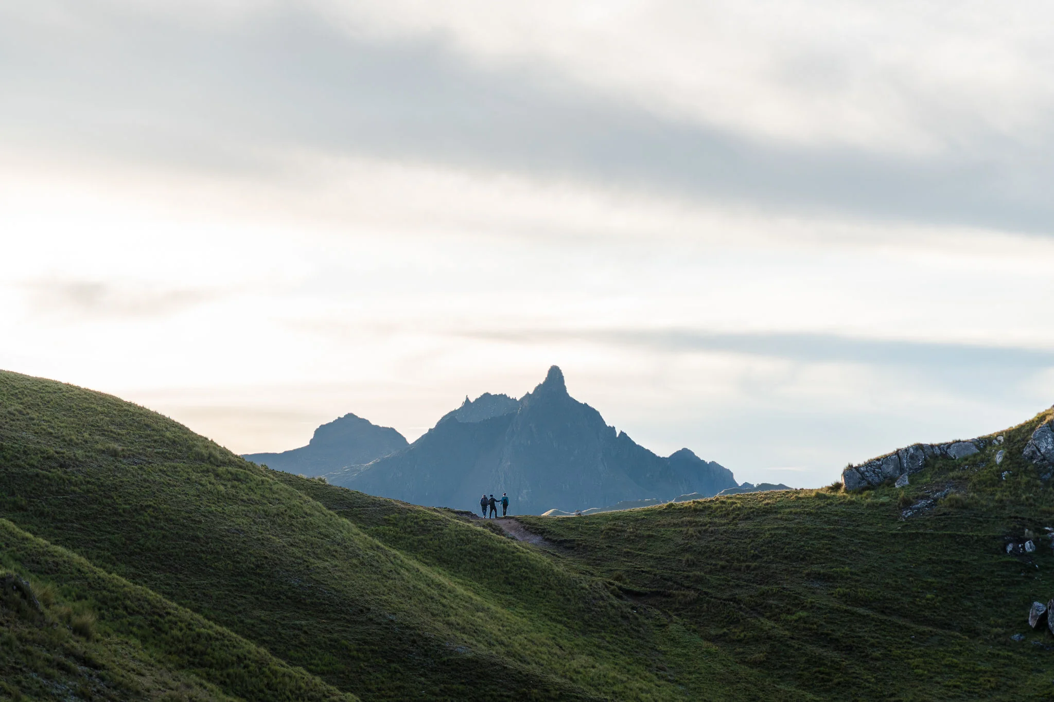 Two hikers silhouetted on a green grassy ridge, a pointed rocky peak rising behind them under a pale sky, Ancascocha, Sacred Valley, Peru