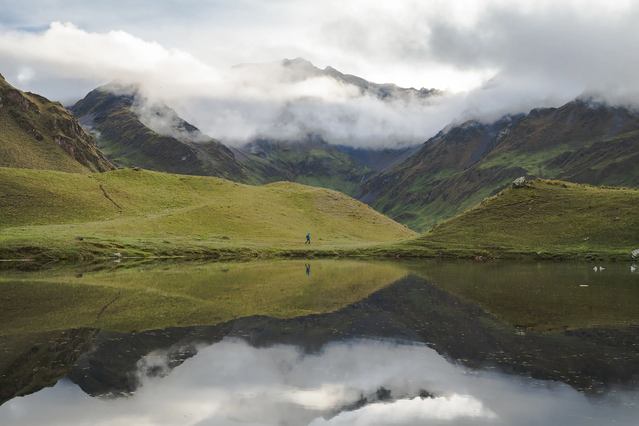 A still lake mirroring green hillsides and low cloud, a lone trekker walking the grassy shore, Ancascocha, Sacred Valley, Peru