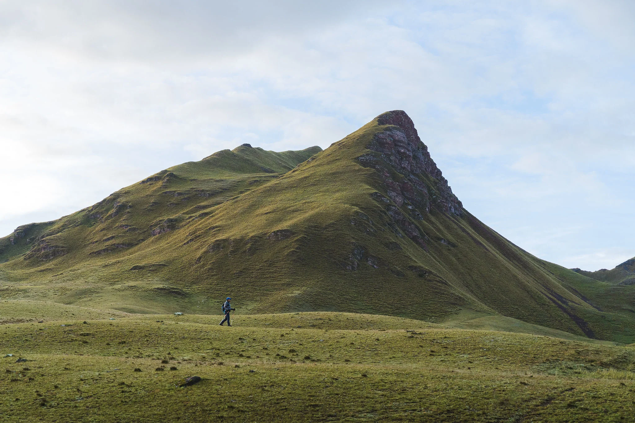 A hiker crossing a wide grass plain in morning light below a steep green peak, Ancascocha, Sacred Valley, Peru