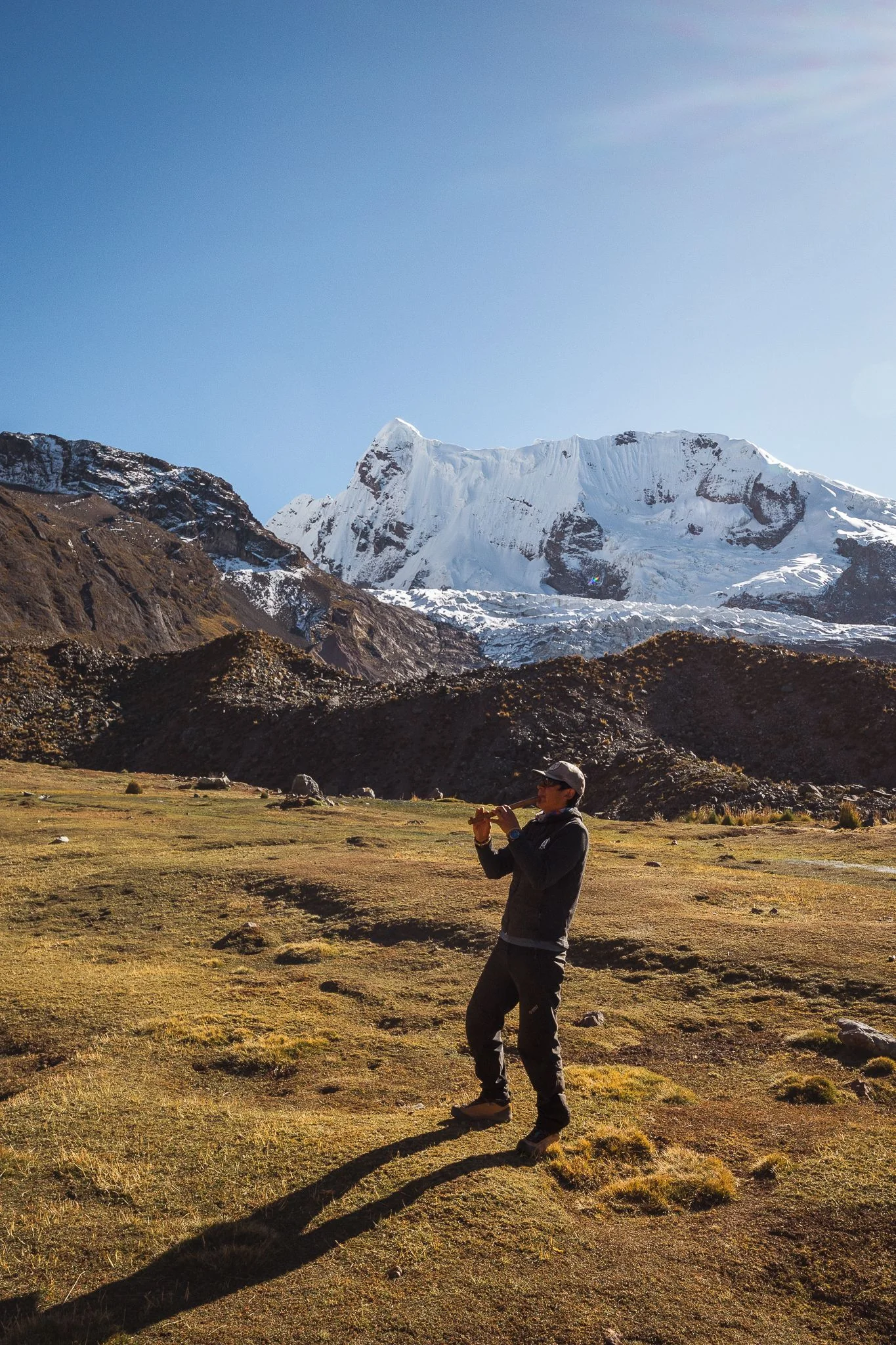 A Peruvian mountain guide in a cap playing the flute on sunlit golden grassland at Nevado Ausangate basecamp, a glaciated snow peak and dark moraine behind him, Cordillera Vilcanota, Peru