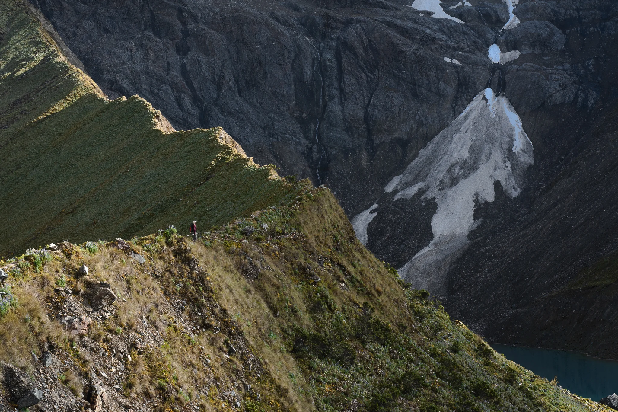 A small hiker on a steep knife-edge grassy ridge beside a sheer dark rock wall with patches of snow and a lake at its foot, Cordillera Vilcabamba, Peru