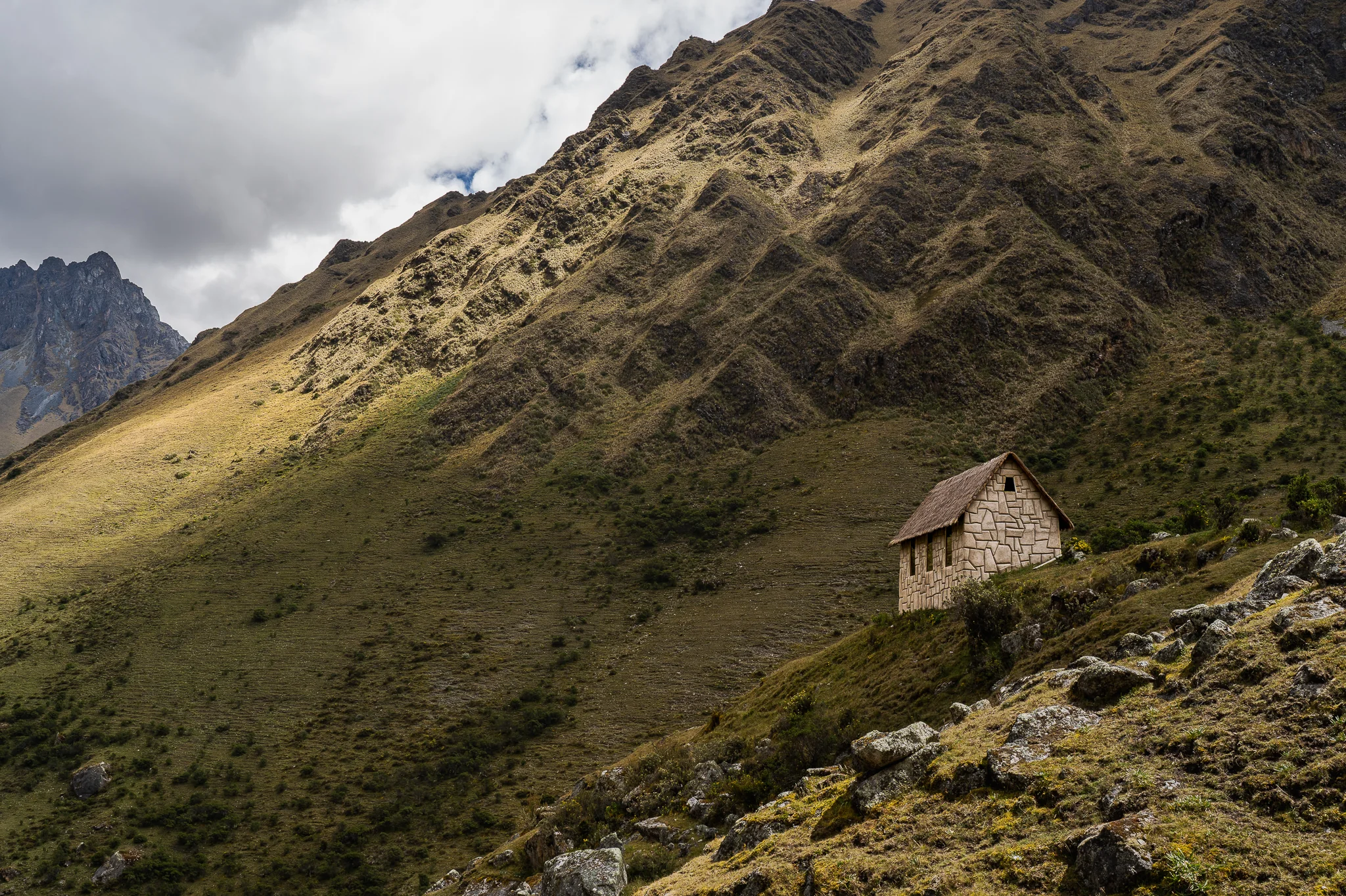 A small stone hut with a pitched roof on a steep green slope, a craggy peak and cloud in the distance, Cordillera Vilcabamba, Peru