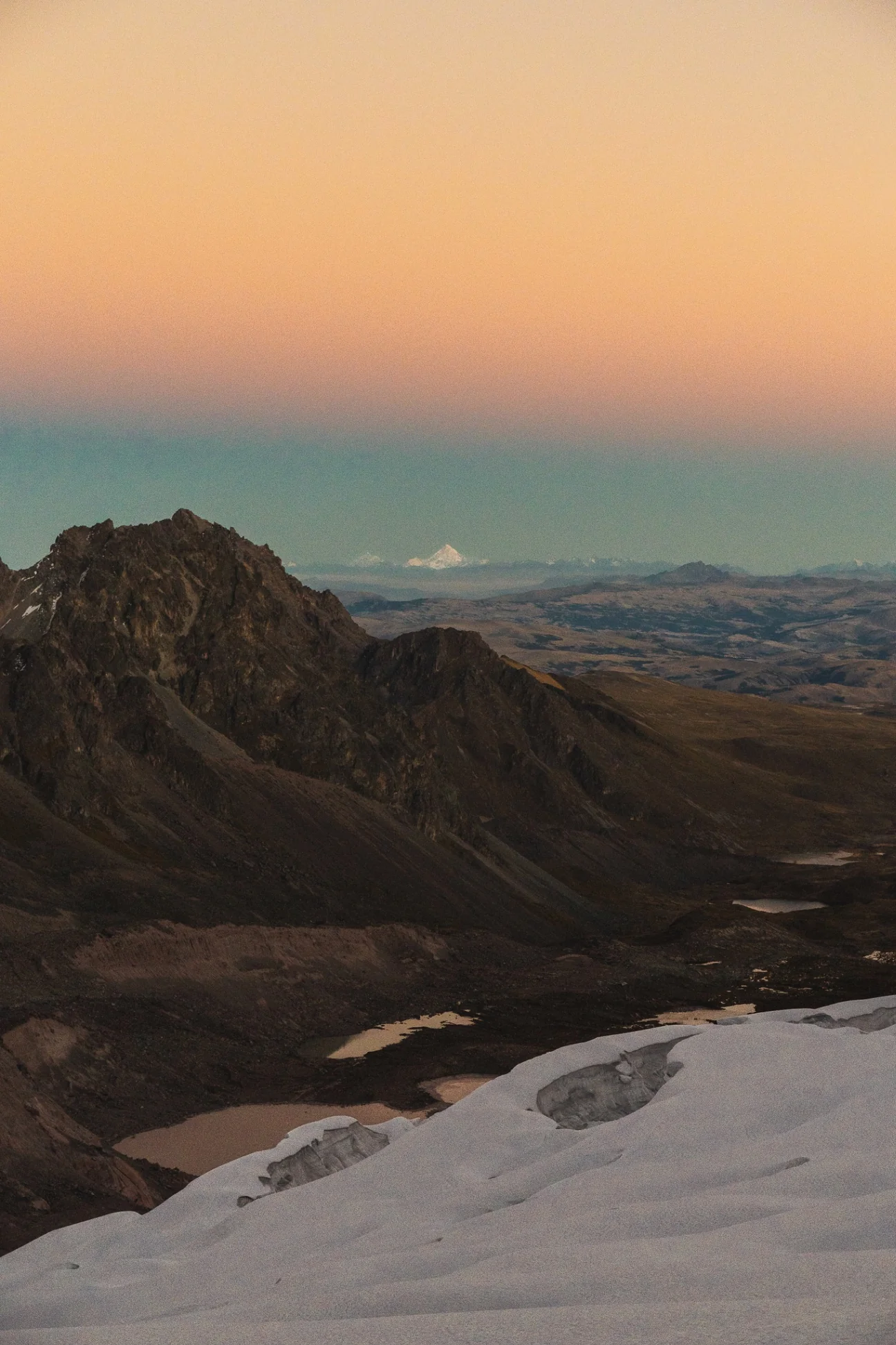 An orange sunrise over a dark rocky peak and a high plateau dotted with small lakes, the northwest glacier of Nevado Campa in the foreground and Nevado Salkantay far off on the horizon, Cordillera Vilcanota, Peru