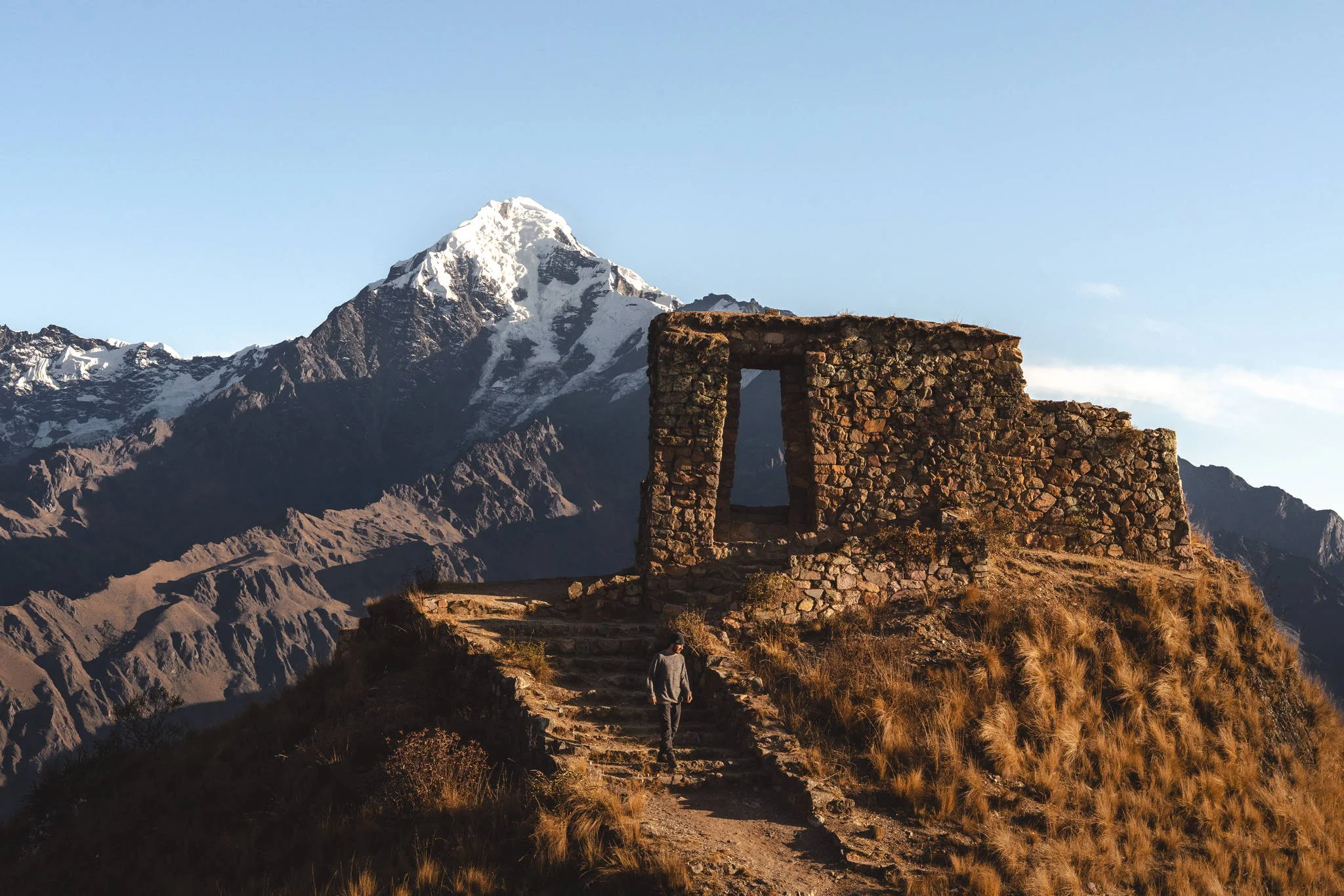 A person walking up the path to the Inca stone gateway of Inti Punku on a ridge, the snow peak of Nevado Verónica rising behind in evening light, Peru