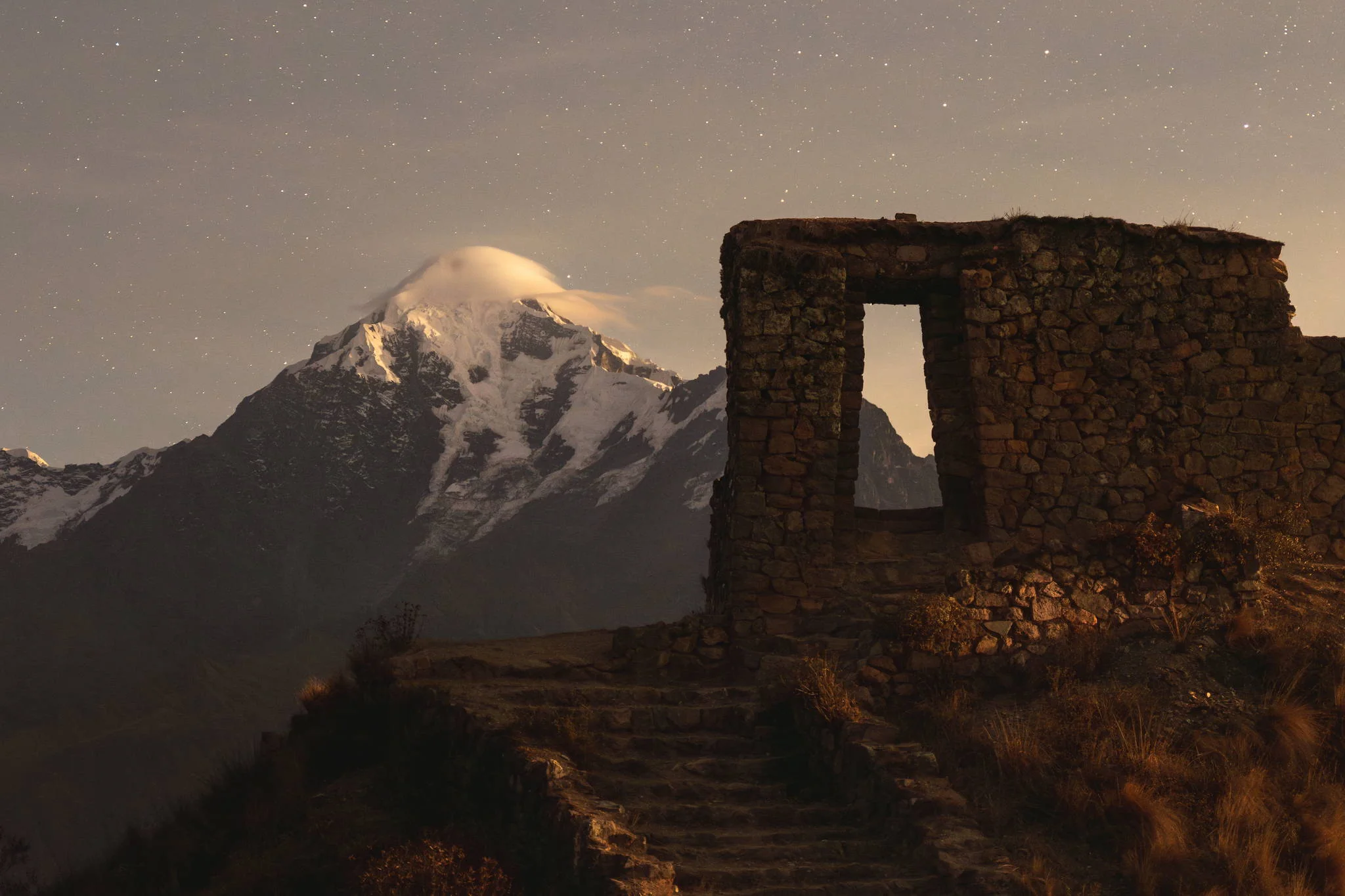 The stone doorway of the Inti Punku ruins at night under a star-filled sky, moonlight on the snow of Nevado Verónica behind, Peru