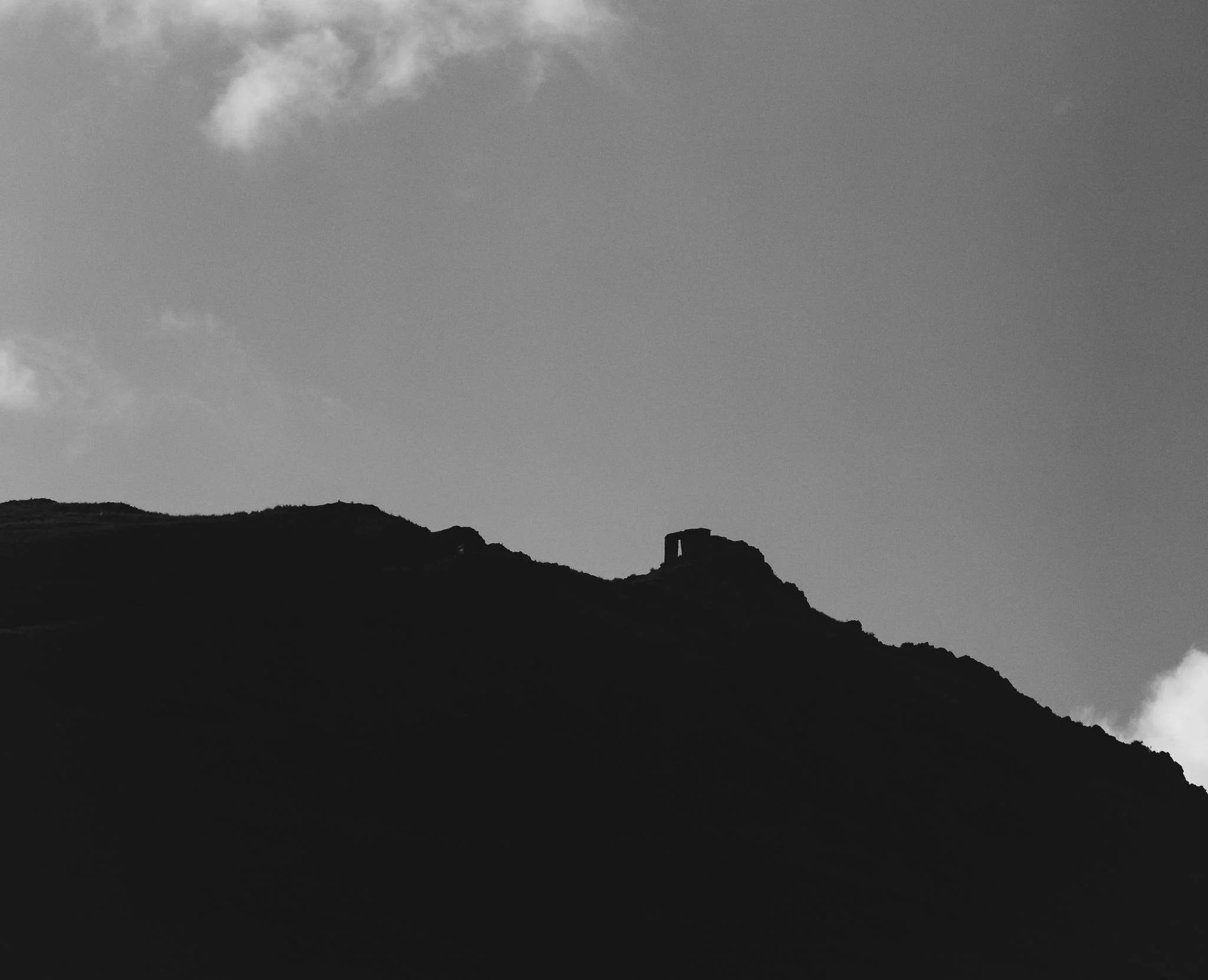 Black-and-white view from the trail below of a ridge in silhouette, the small stone ruin of Inti Punku standing on its crest, Peru