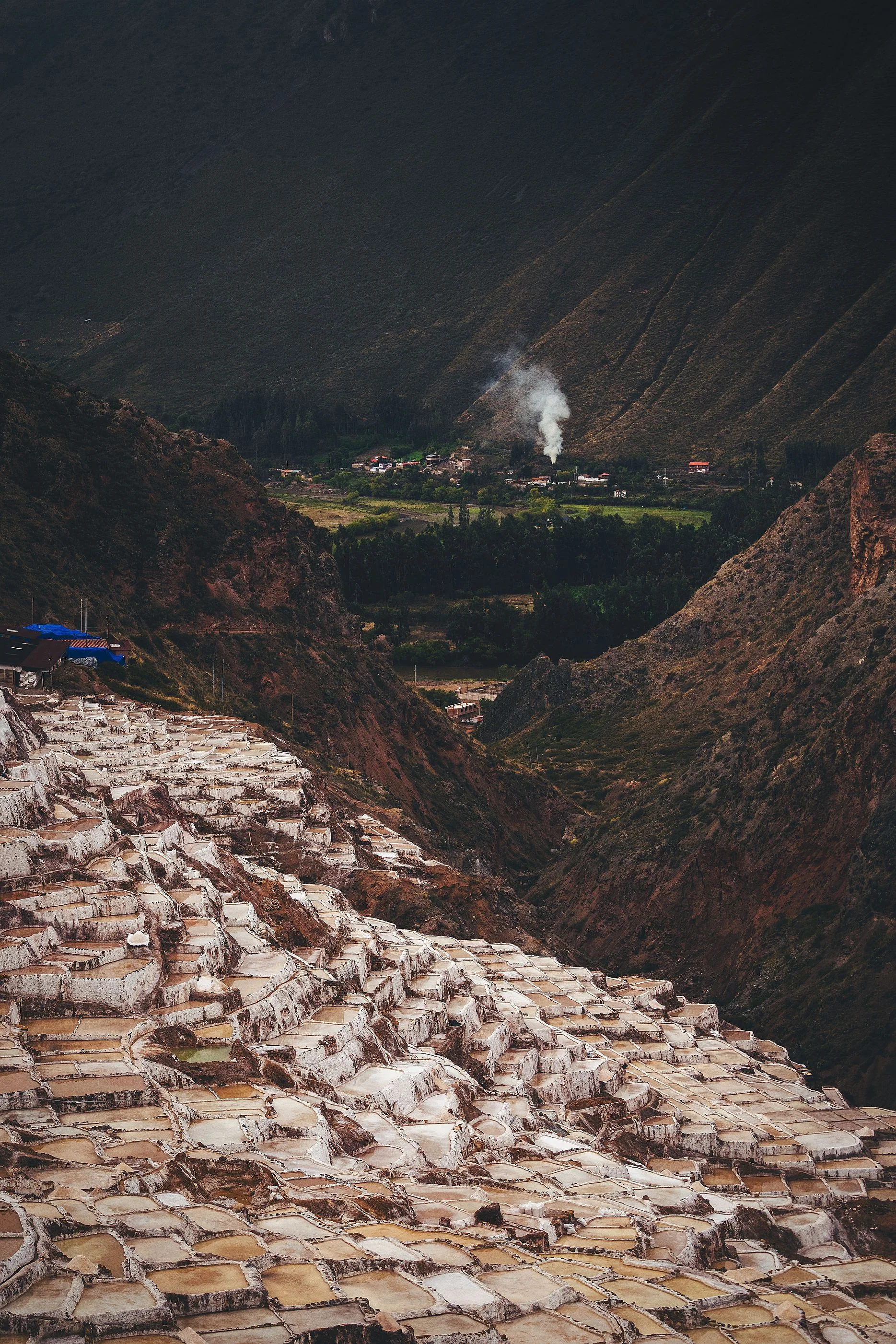 Hundreds of terraced salt pans filling a steep ravine, with a village and green fields on the valley floor below, Maras, Sacred Valley, Peru