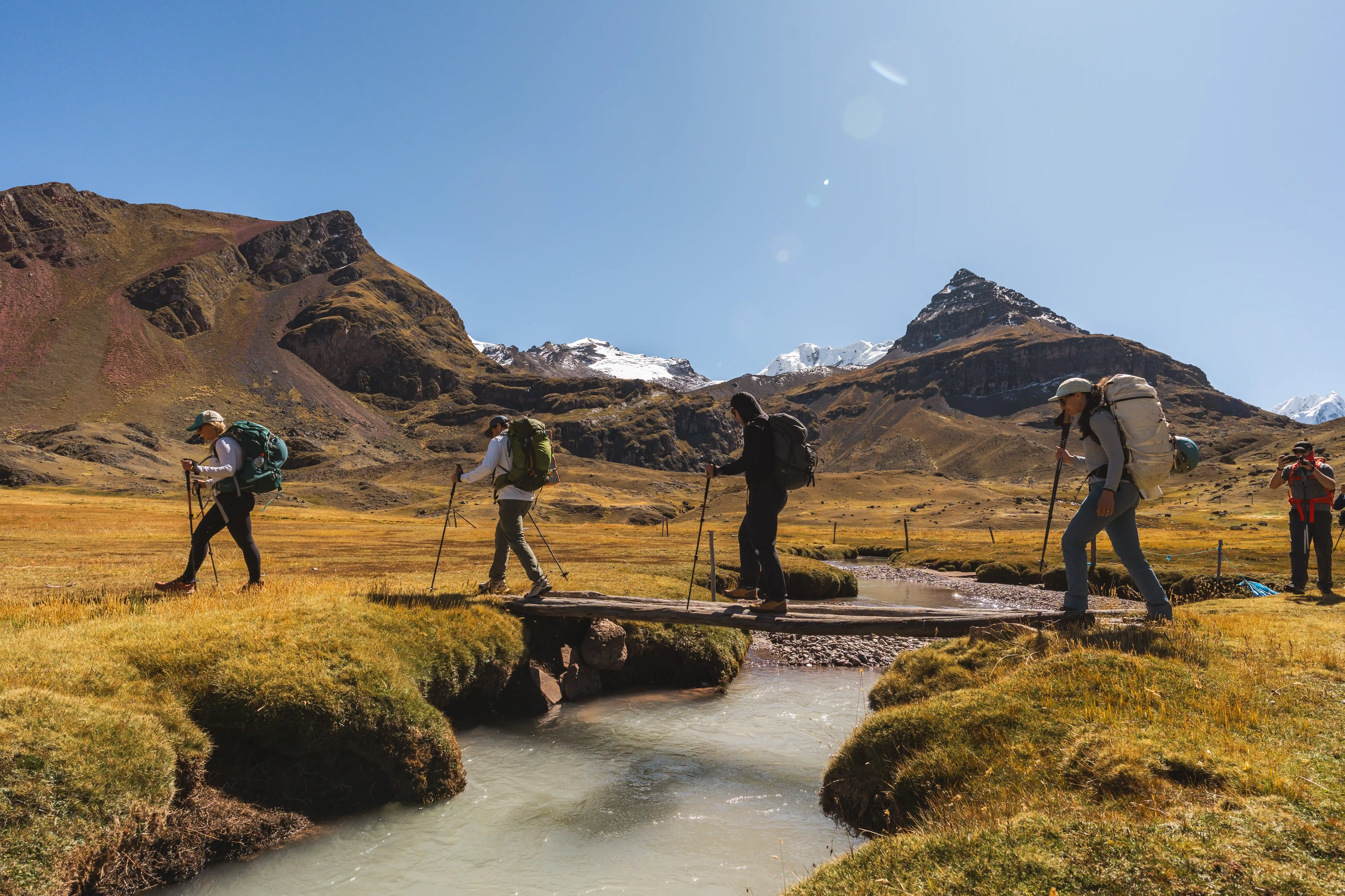 Four trekkers with large packs crossing a stream on a narrow plank bridge, golden grassland and snow-dusted peaks beyond, on the way to Ausangate base camp, Peru