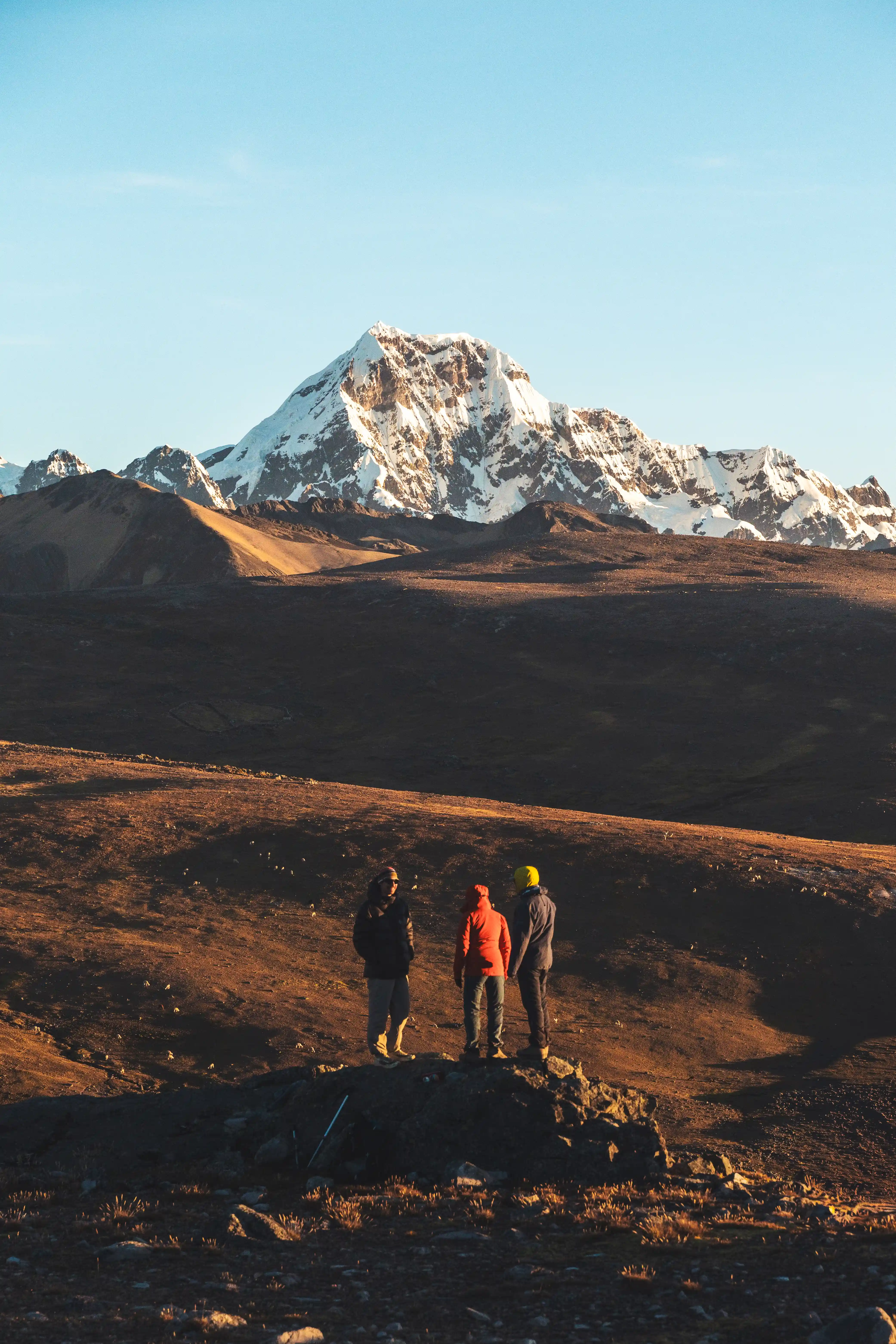 Three climbers standing on a rock outcrop at sunset, the snow-covered massif of Nevado Ausangate rising across the plain behind them, Peru