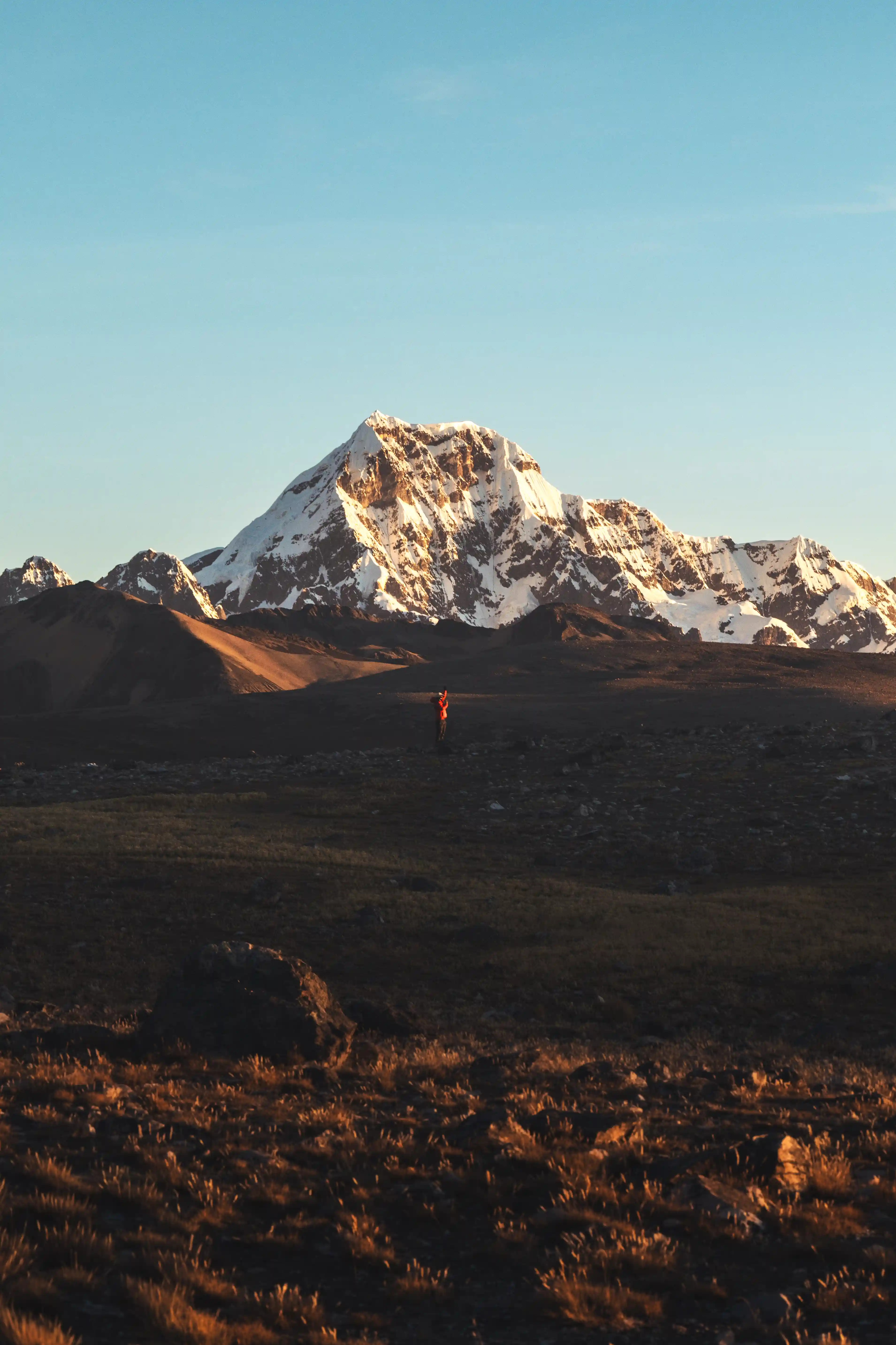 A climber in a red jacket alone on a dark plain at sunset, the snow-covered summit of Nevado Ausangate catching the last light behind, Peru