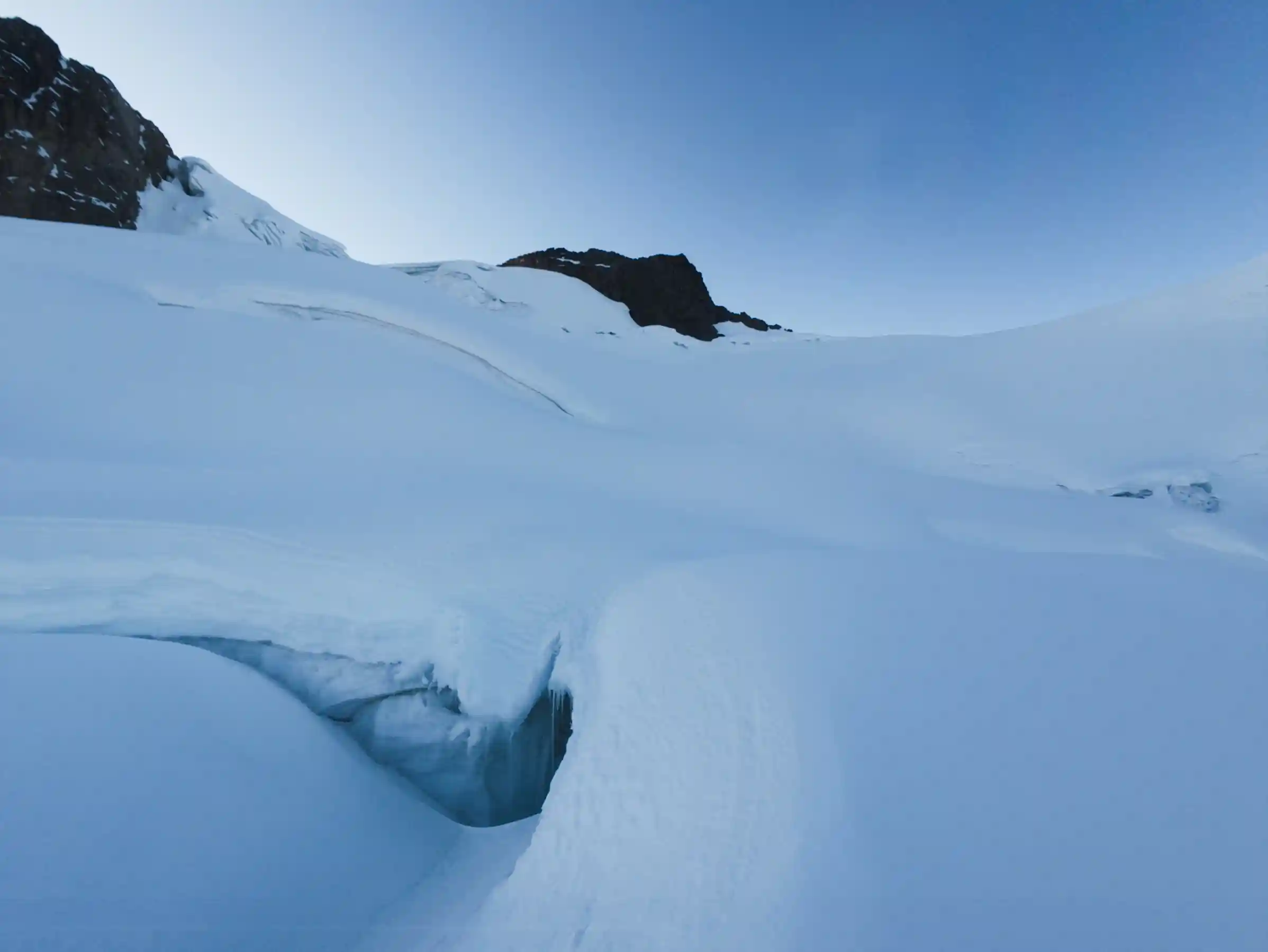 A climber's view up snow slopes past the open lip of a crevasse towards dark summit rocks, northwest side of Nevado Sirijuani, Peru