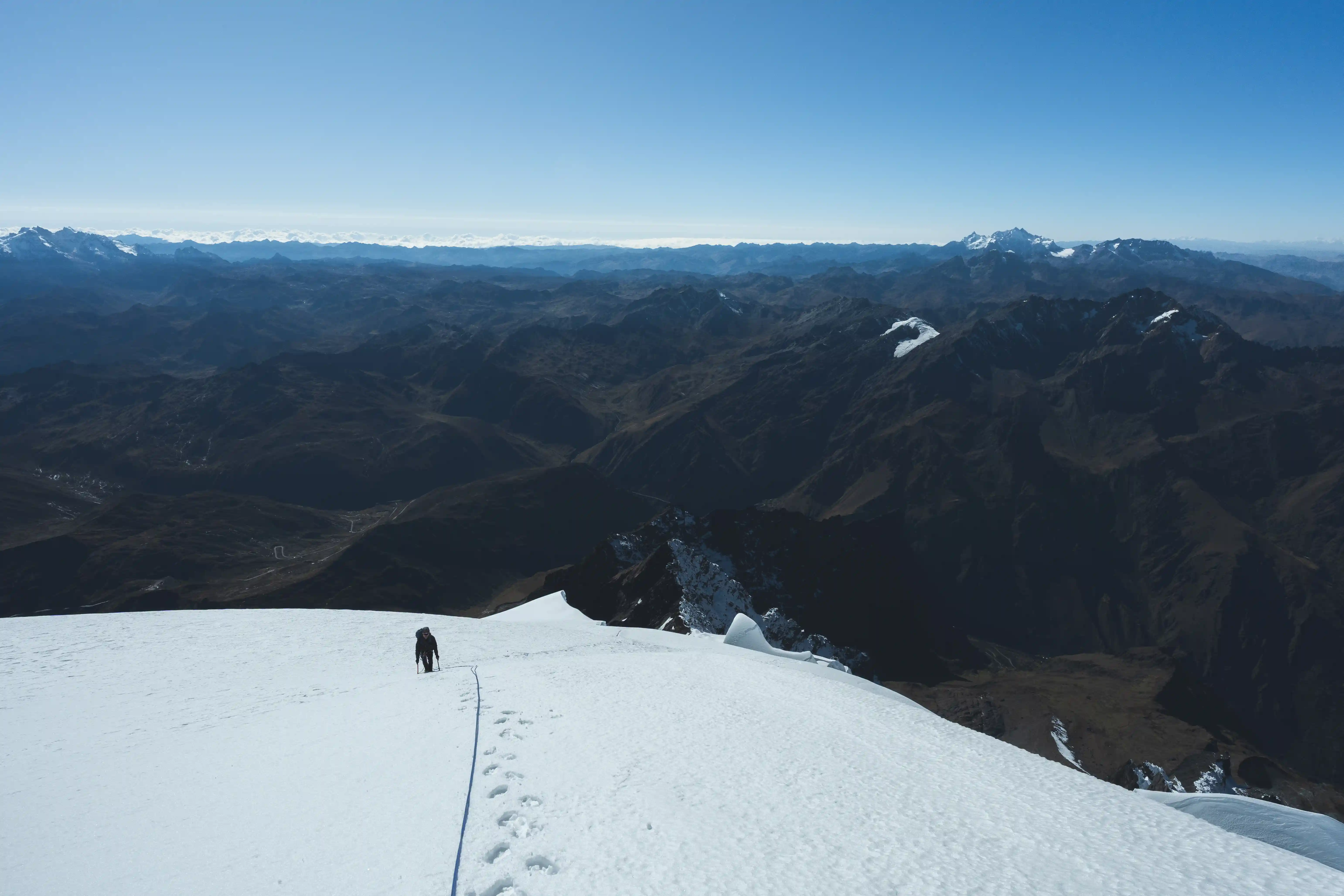 A climber walking the broad snow dome near the summit of Nevado Verónica, range after range spreading out far below, Peru