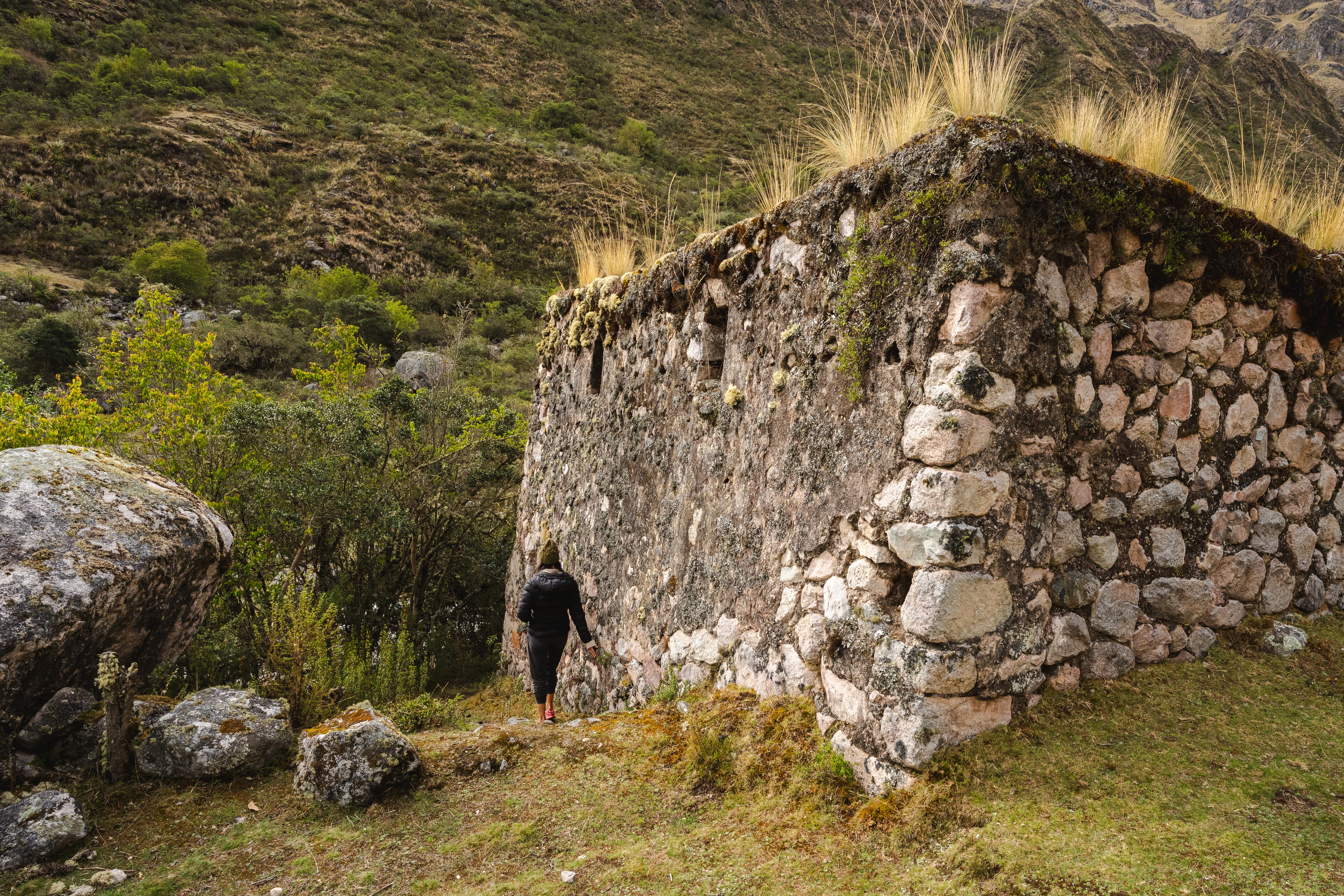 A person standing at the corner of a tall dry-stone ruin topped with grass, boulders and scrub around it, Pumahuanca valley, Peru