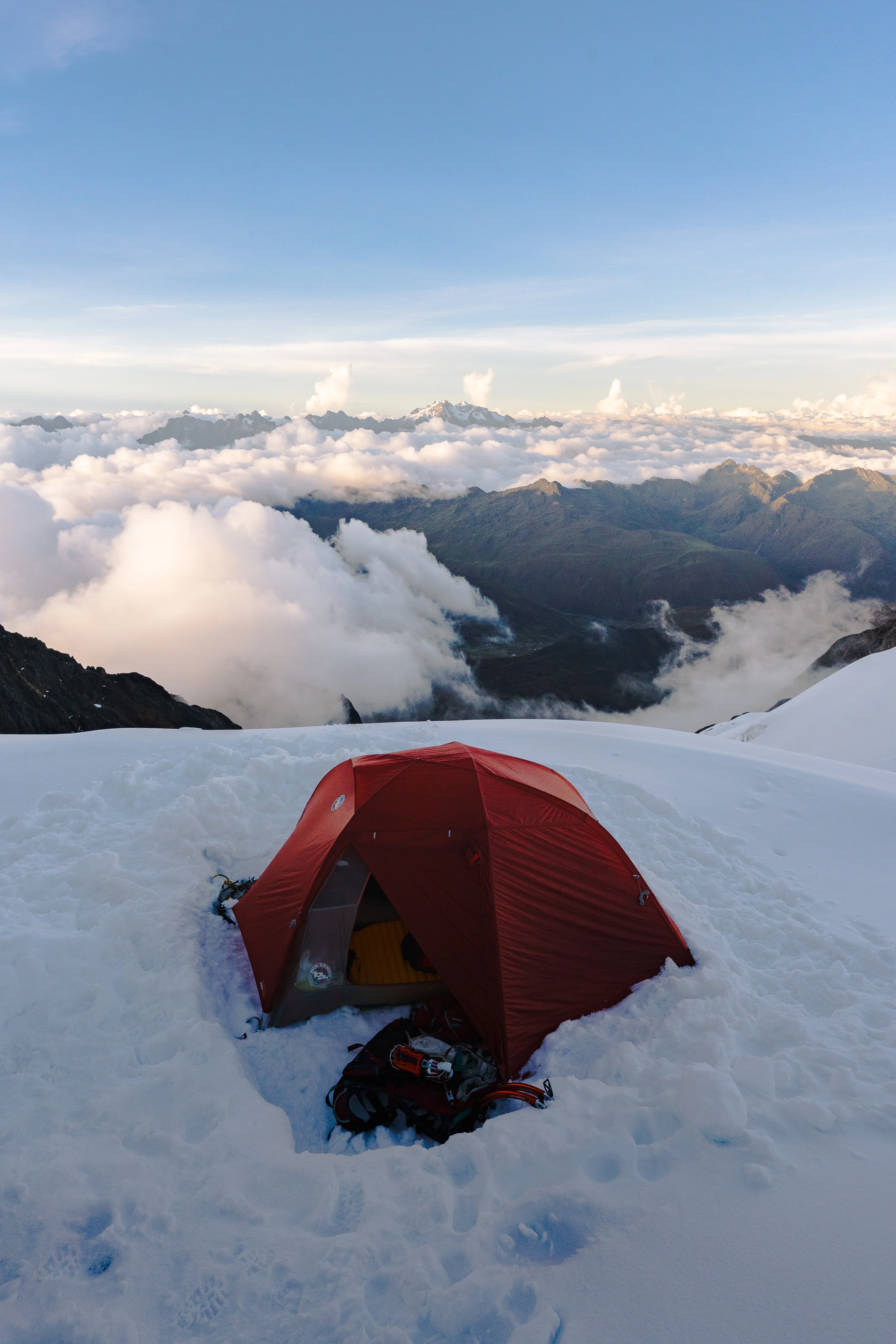 A red dome tent pitched in a hollow dug into snow, a backpack at its entrance and cloud filling the valleys below, Cordillera Urubamba, Peru