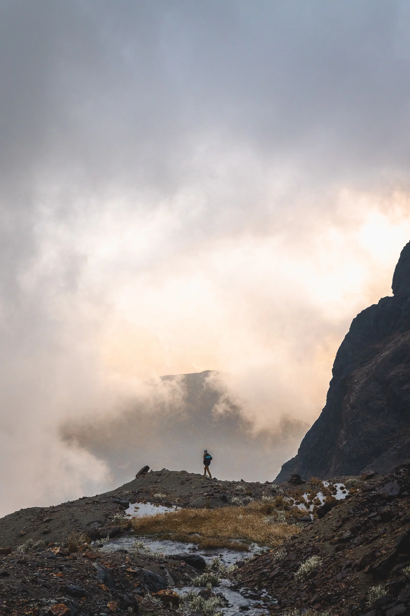 A climber standing on a rocky shoulder at sunset as cloud pours past, a dark rock tower behind, Nevado Verónica base camp, Peru