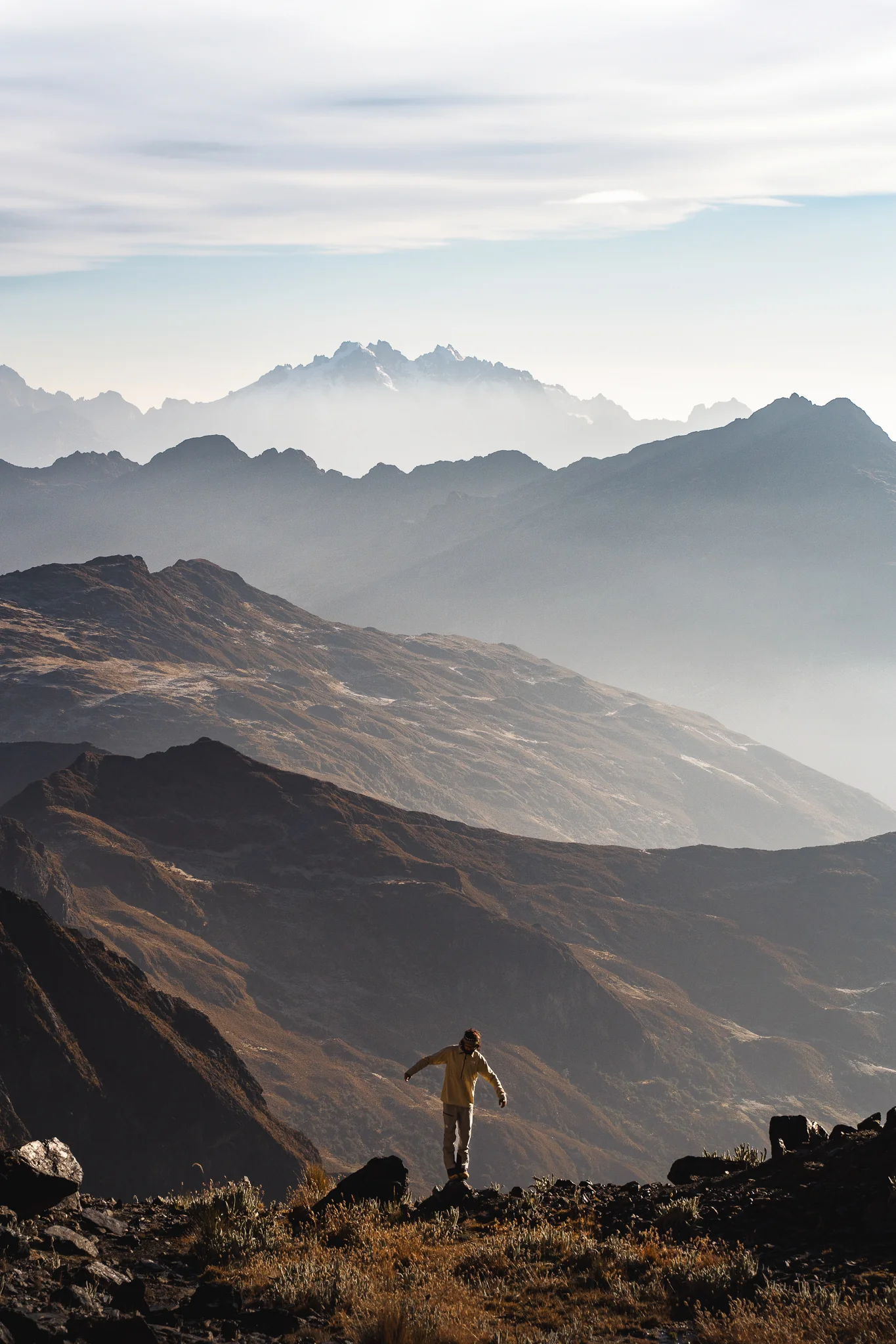 A climber balancing with arms outstretched on a rocky edge at dusk near Nevado Verónica basecamp, layers of hazy ridges and the jagged Nevados Terihuay in the distance, Cordillera Urubamba, Peru