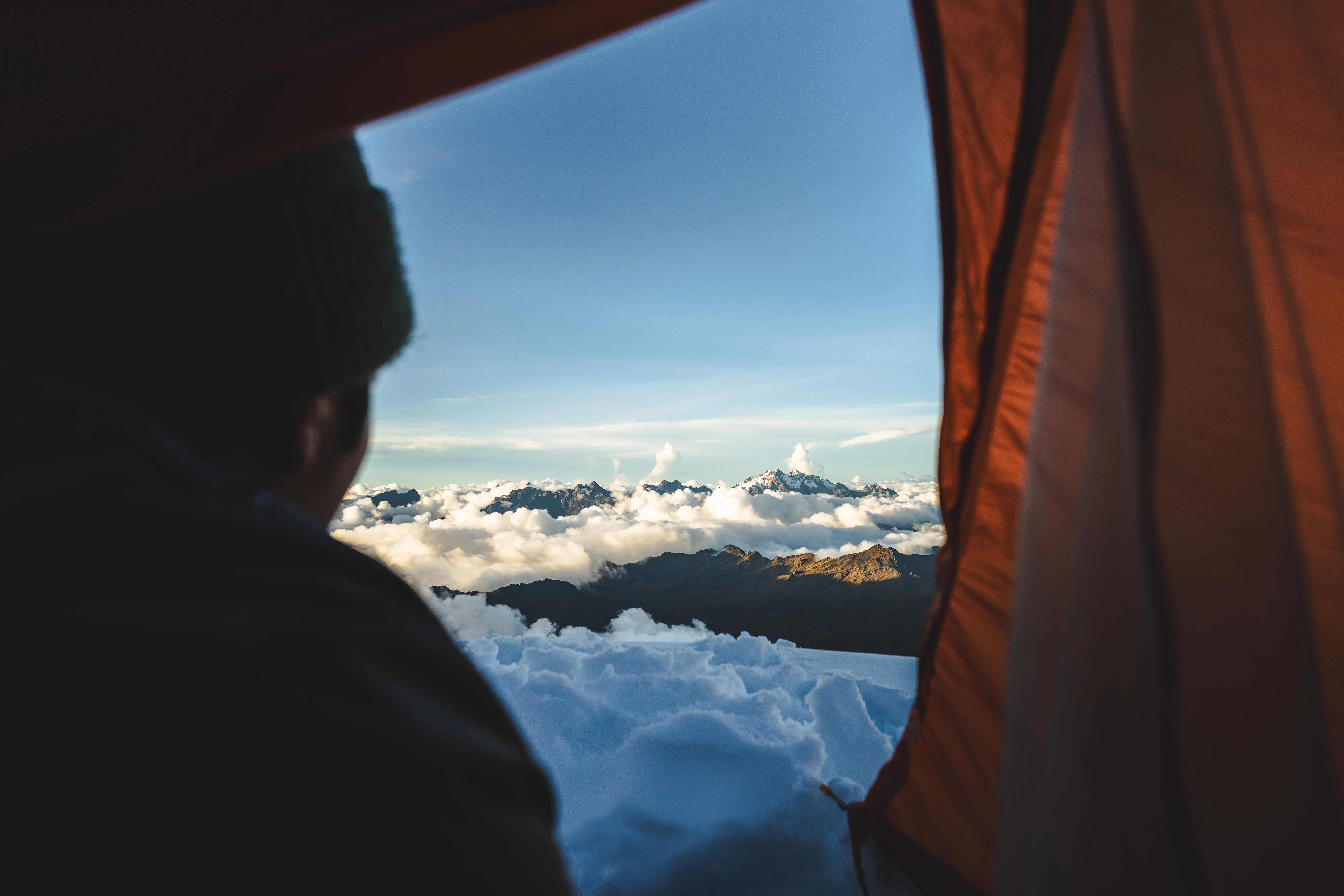 A climber in a beanie looking out of an orange tent doorway at a sea of cloud and distant snow peaks, Cordillera Urubamba, Peru