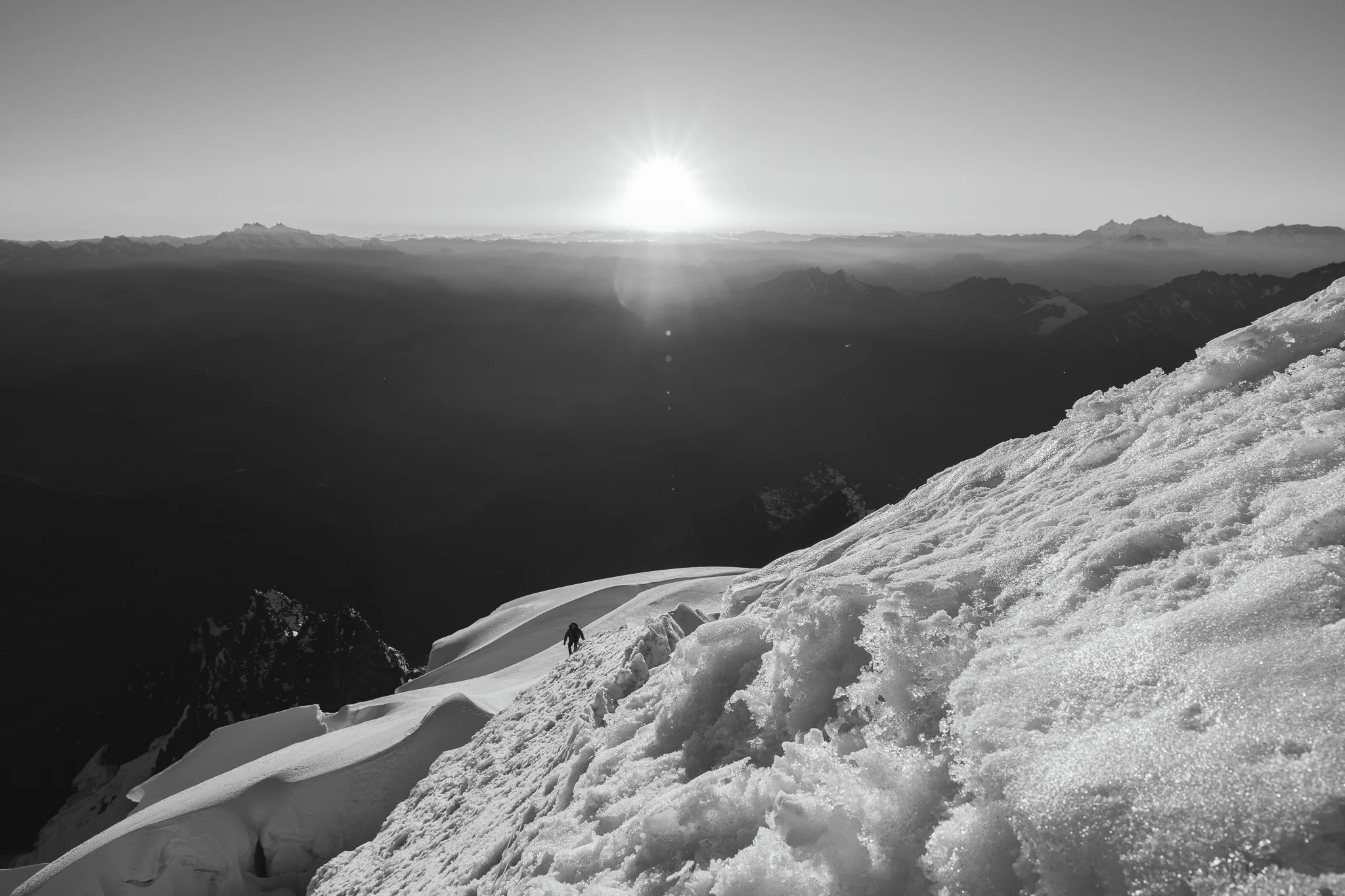 Black-and-white photo of a climber on the final snow slopes of Nevado Verónica, the sun low over ridges and cloud beyond, Peru